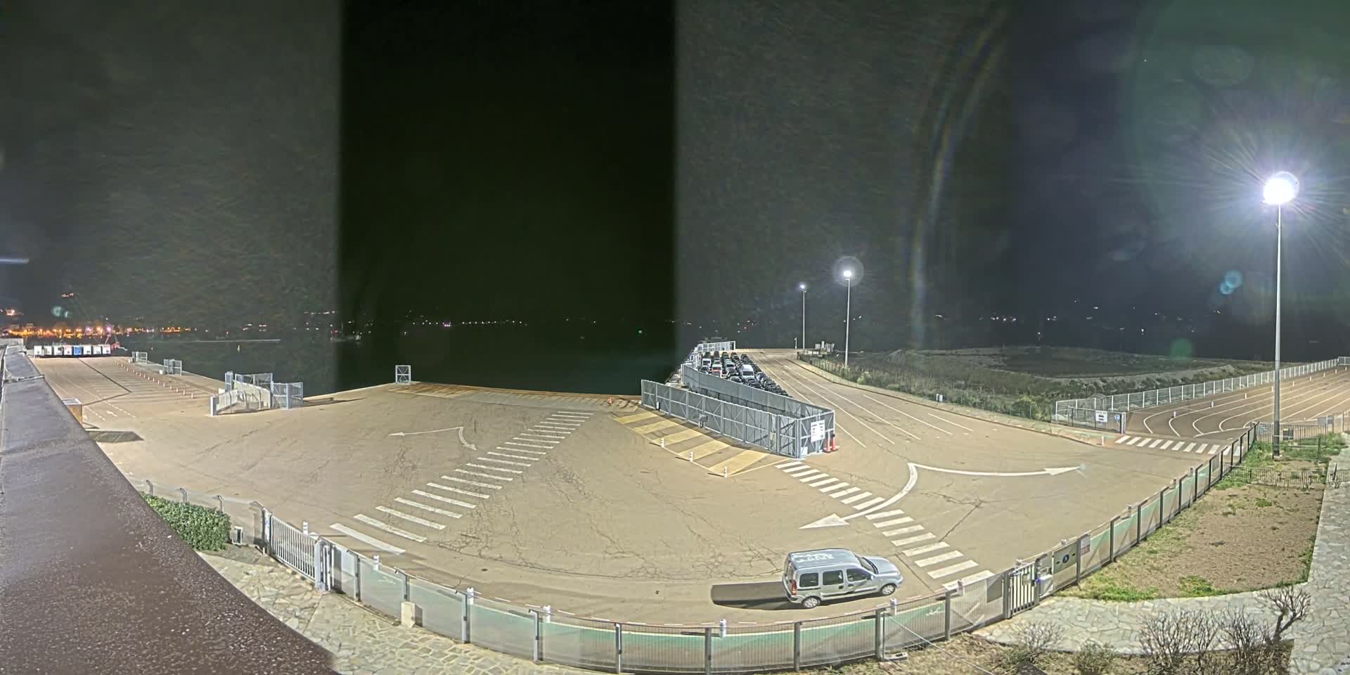 An empty, paved area with crosswalks and a single vehicle is shown at night near a body of water, under bright artificial lights.