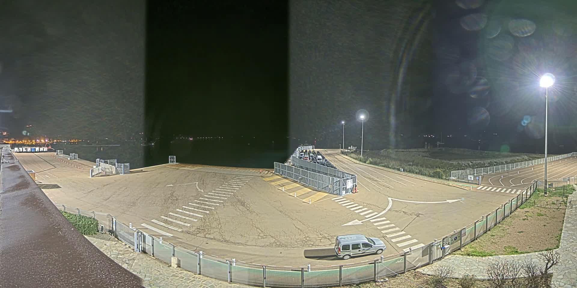 An empty, nighttime ferry terminal with a single vehicle parked near a fenced-off area containing a line of other cars, overlooking calm water under bright artificial lights.
