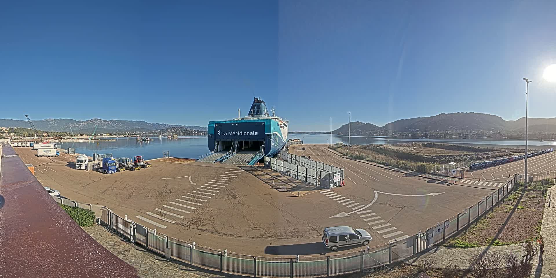 A teal and white ferry is docked at a port on a sunny day, with mountains in the background and several vehicles parked nearby.