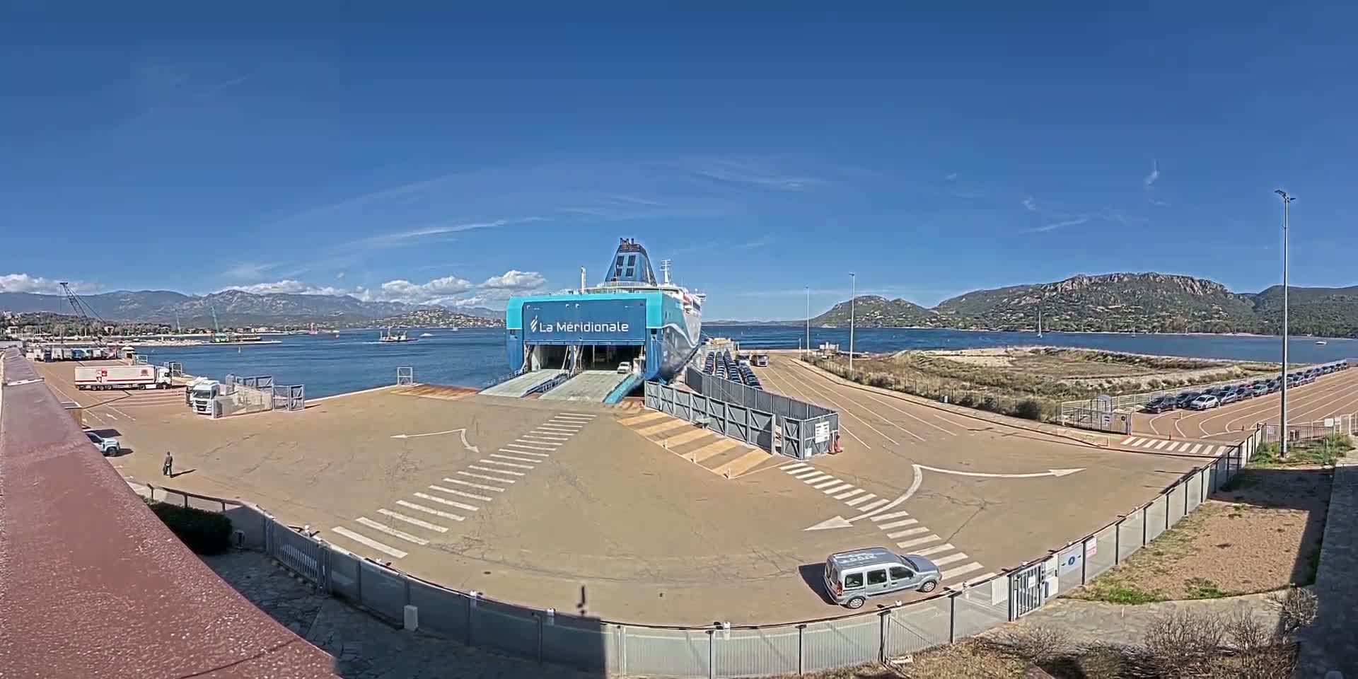 A large ferry is docked at a port on a sunny day, with mountains and calm water in the background; several vehicles are parked nearby.