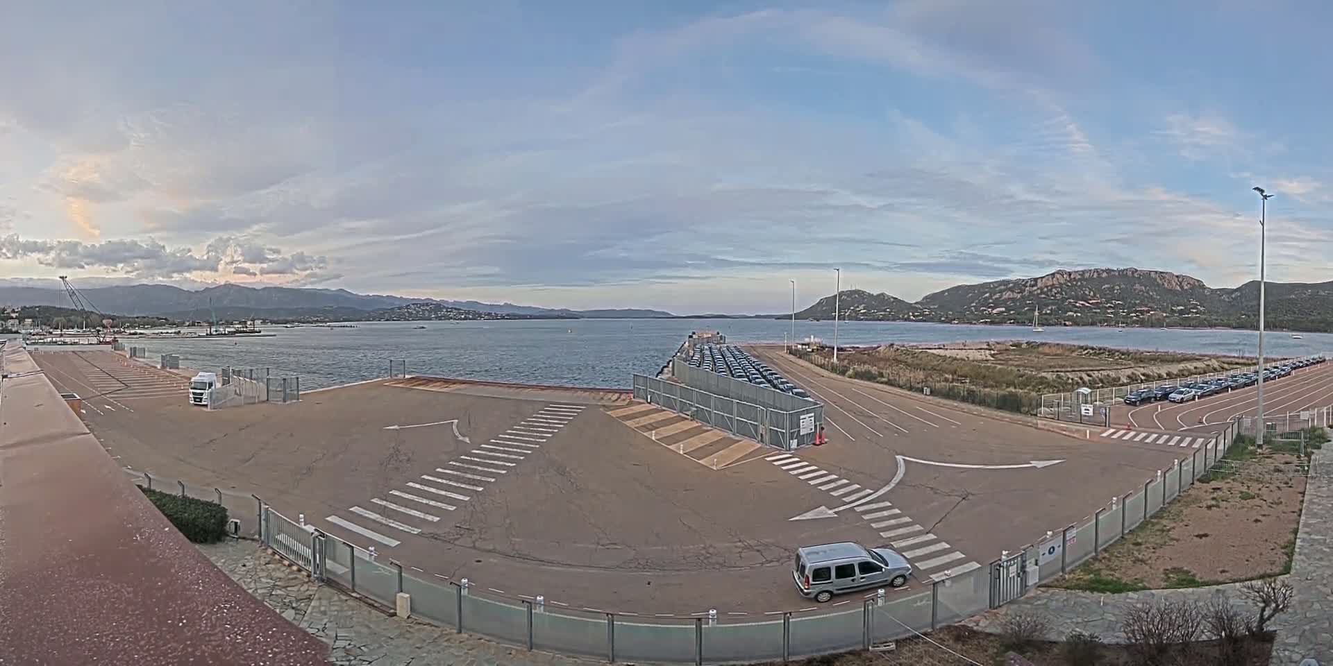 An empty paved area with crosswalks and parked cars near a body of water under a partly cloudy sky, mountains in the background.