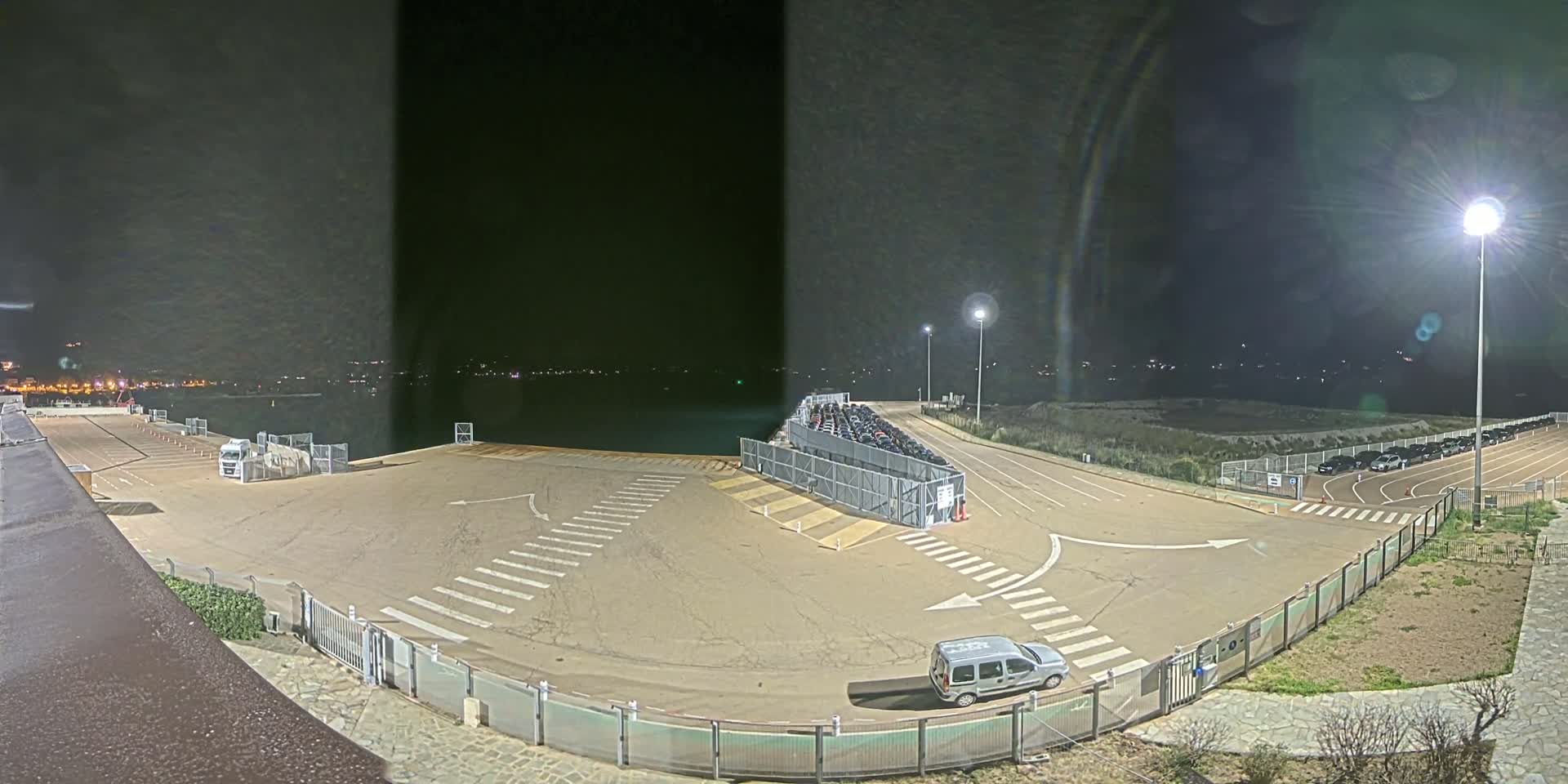 An empty, nighttime parking lot next to a body of water, with several cars parked in a fenced area and one vehicle in the lot, under bright artificial light.