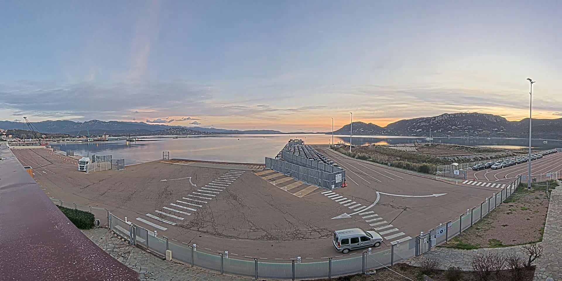 A mostly empty paved lot with a few vehicles, bordered by a calm body of water with mountains in the background under a partly cloudy sunset sky.