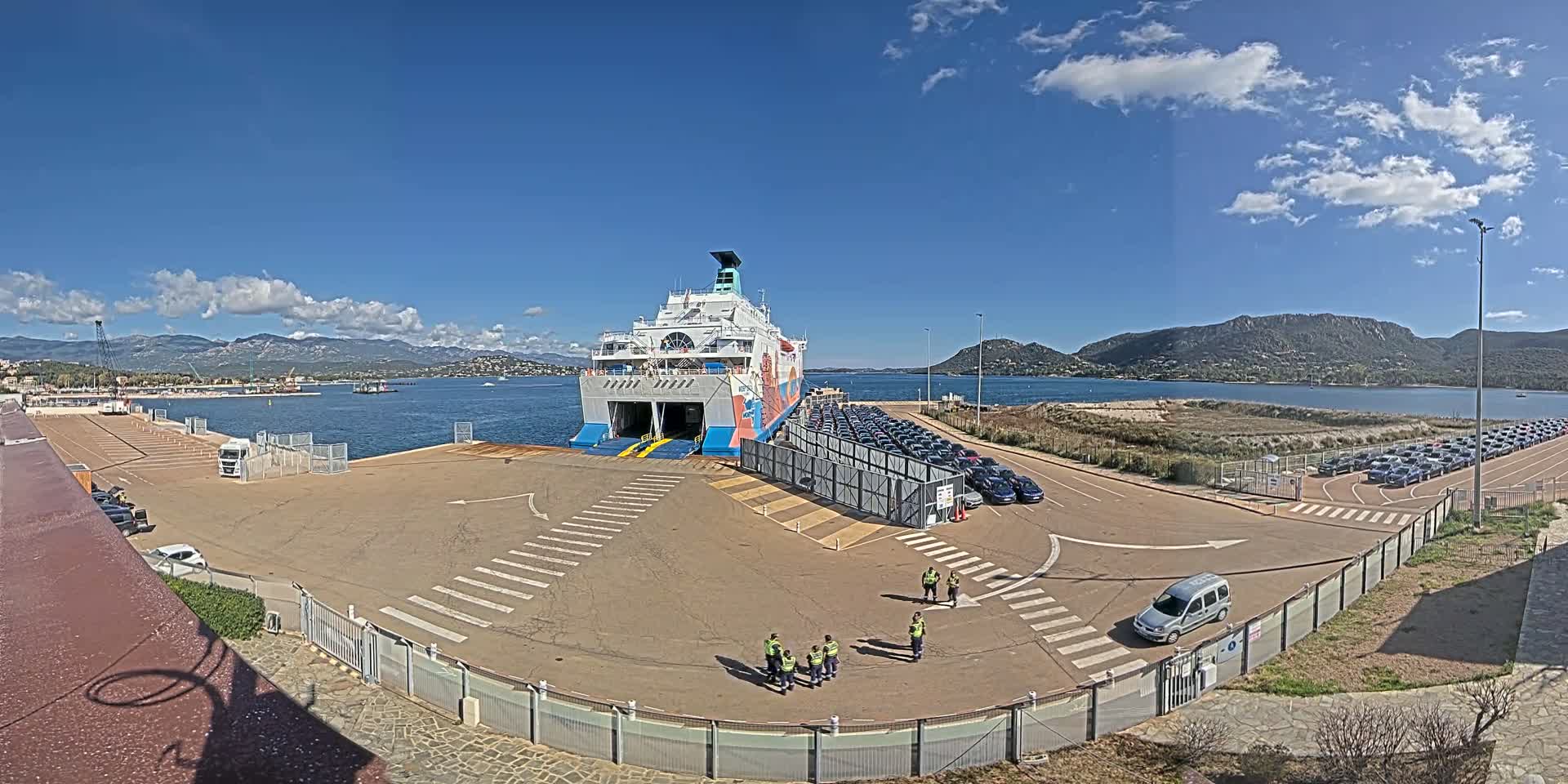 A large ferry is docked at a port on a sunny day, with numerous cars parked nearby and a few people standing on the dock.
