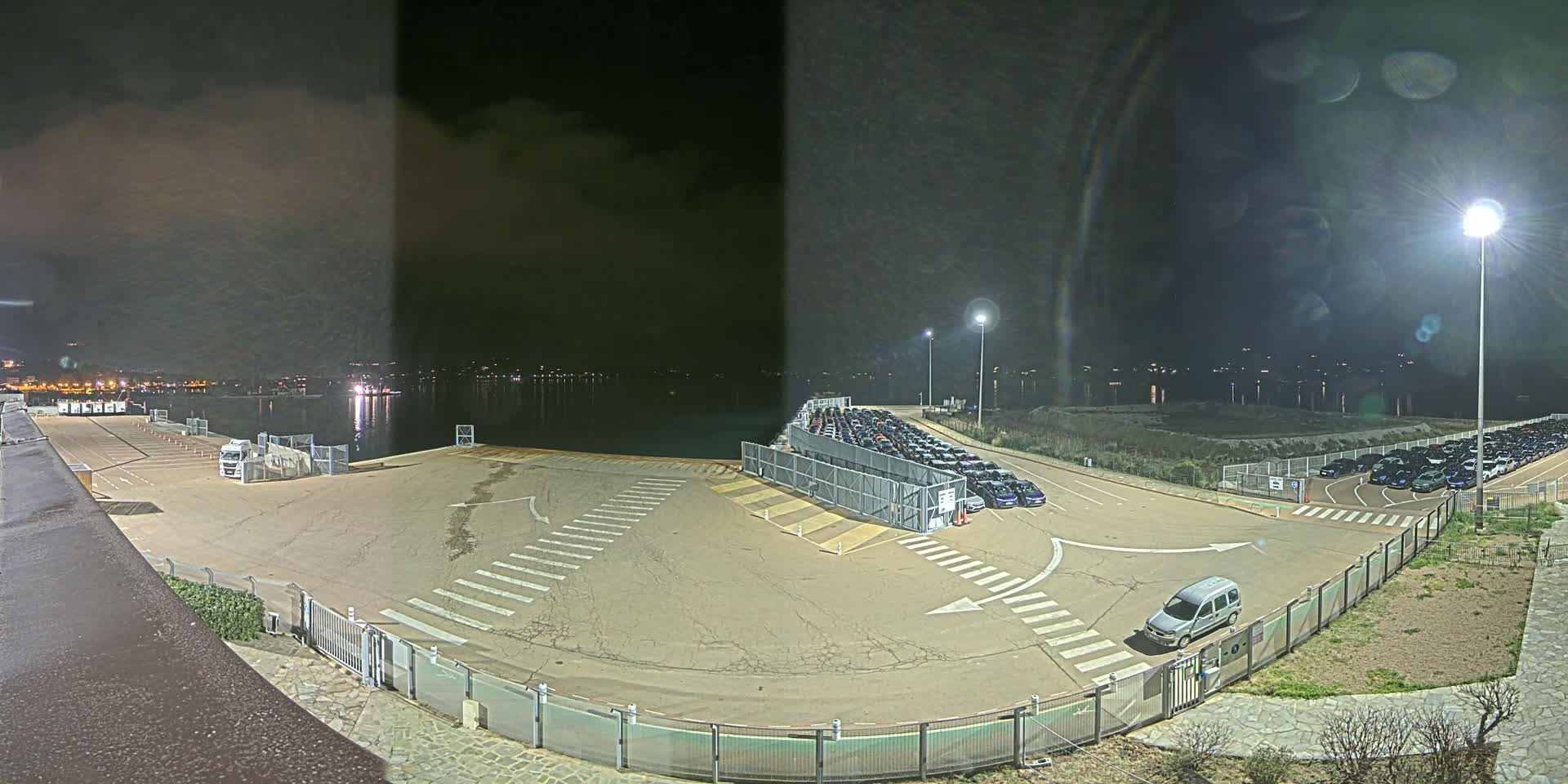 A nighttime view of a mostly empty parking lot near a body of water, with rows of parked cars visible in the distance under bright artificial lights.