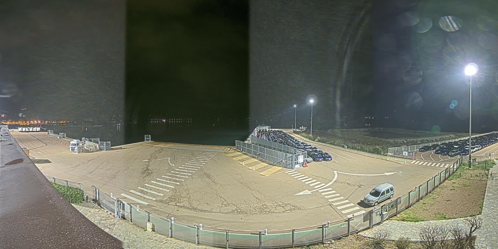A nighttime view of a port area with a large parking lot filled with cars, a body of water in the background, and a single van near a gate.