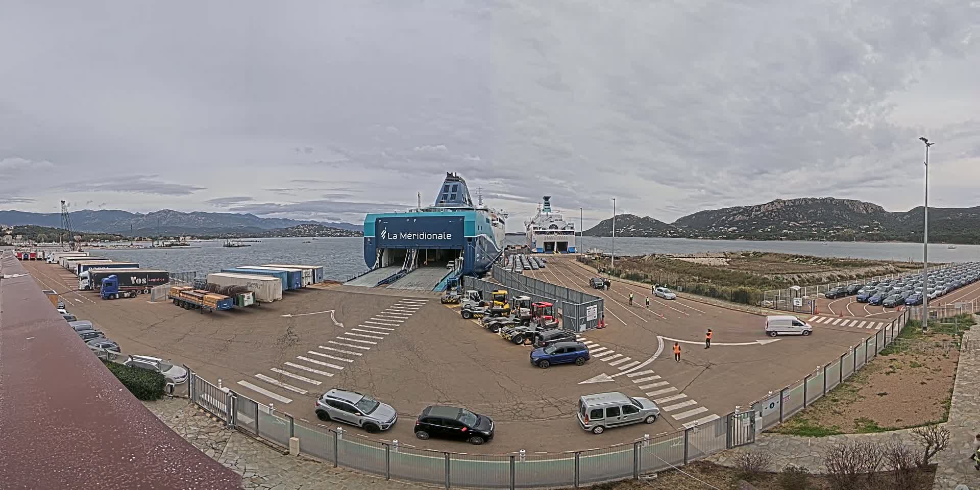 A ferry is docked at a port on an overcast day, with numerous cars and trucks nearby, and mountains visible in the background.