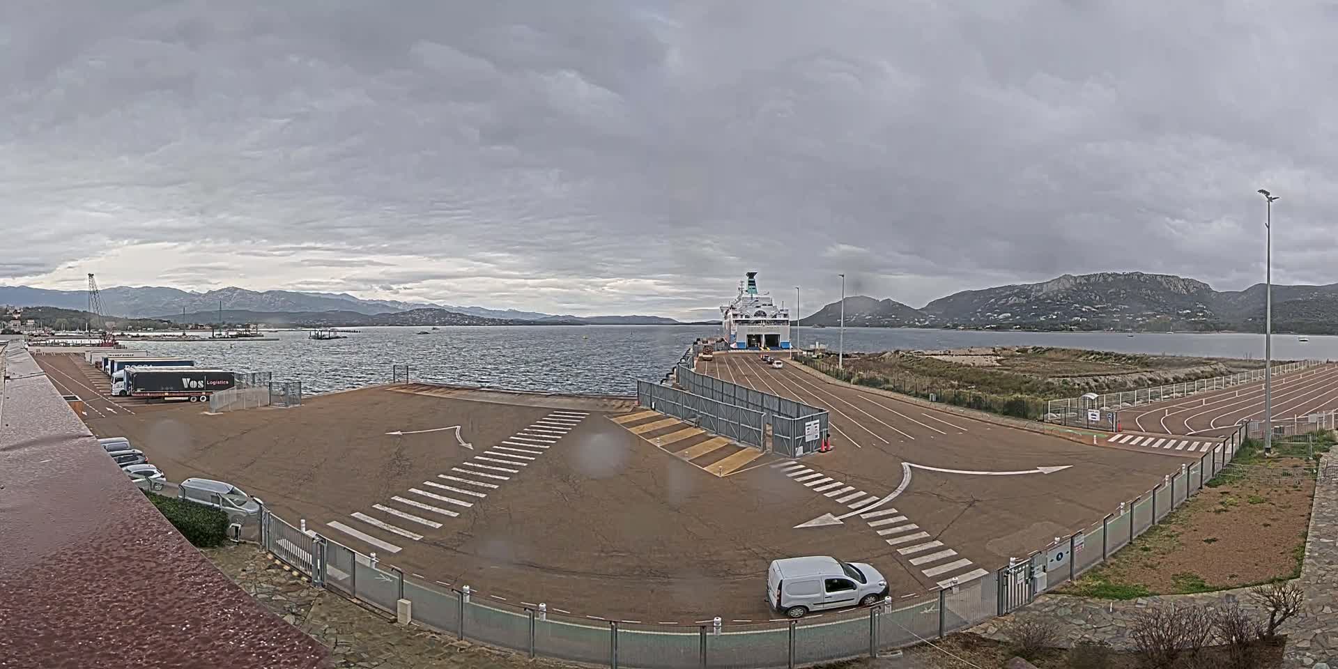 A ferry is docked at a port under a cloudy sky, with vehicles parked in the surrounding area.