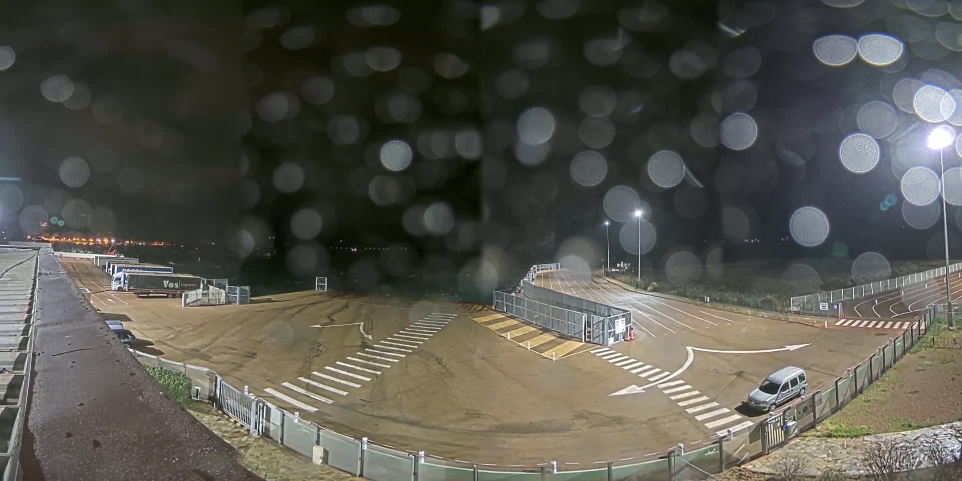 A nighttime, rain-soaked view of a mostly empty parking lot with crosswalks and a few parked vehicles, including a van and several semi-trucks.