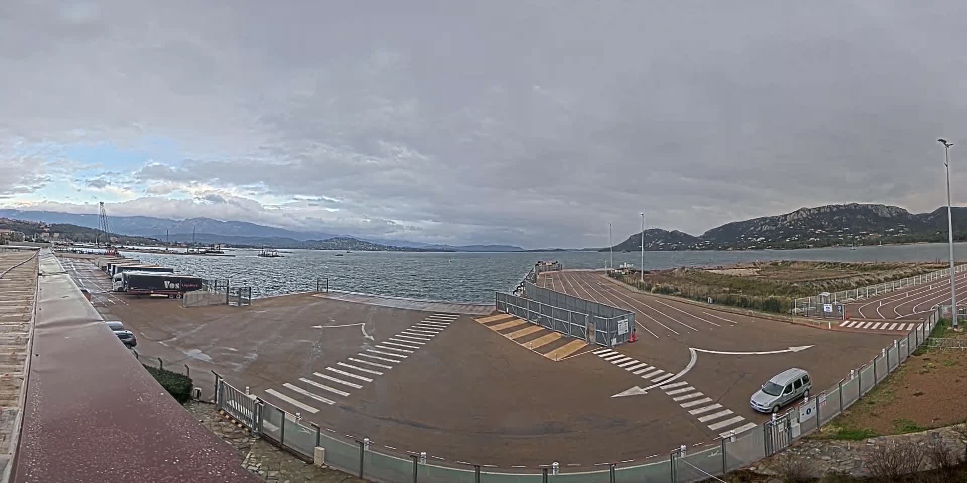 A waterfront parking lot with a few vehicles, under a cloudy sky, overlooks a calm body of water and distant mountains.