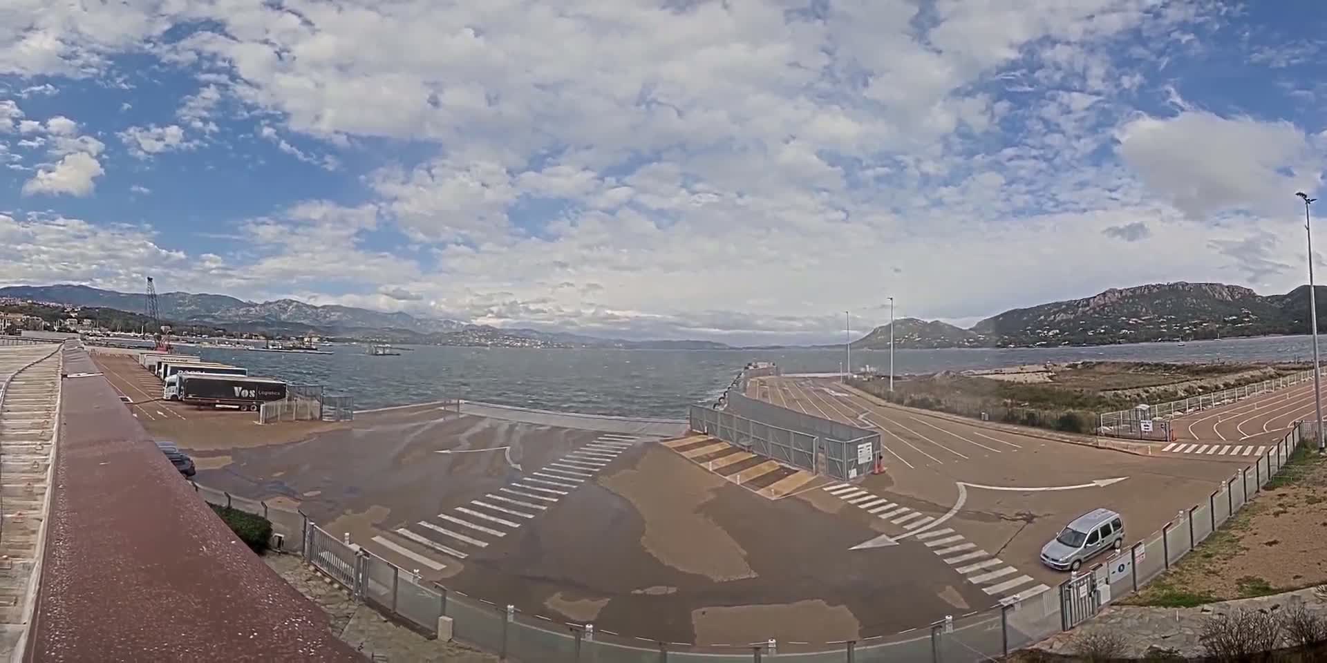 A ferry terminal with several trucks and one car is situated near a body of water under a partly cloudy sky.