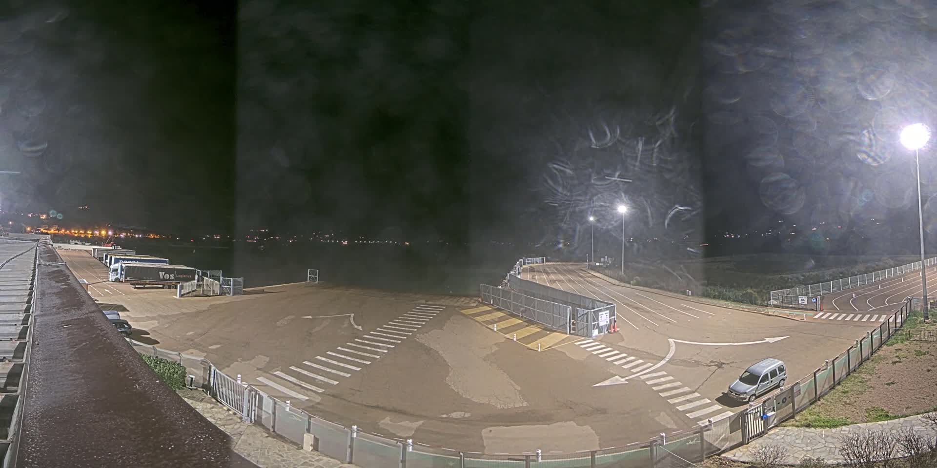 A nighttime view of an empty paved lot with some parked trucks and a single car, under a hazy sky illuminated by several bright lights.