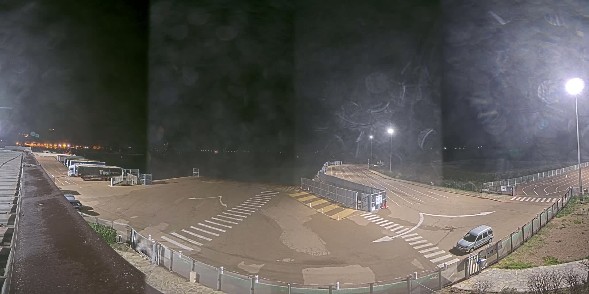 An empty, asphalt lot at night, with a few trucks parked and a single car, under a hazy, dark sky illuminated by artificial lights.