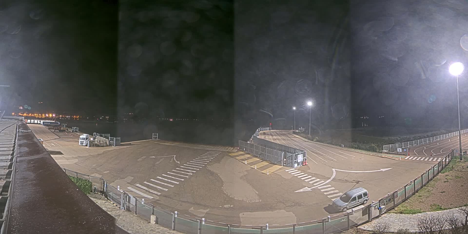A nighttime view of an empty, asphalt roadway with crosswalks and a few vehicles, bordered by fences and floodlights, overlooking a dark body of water.