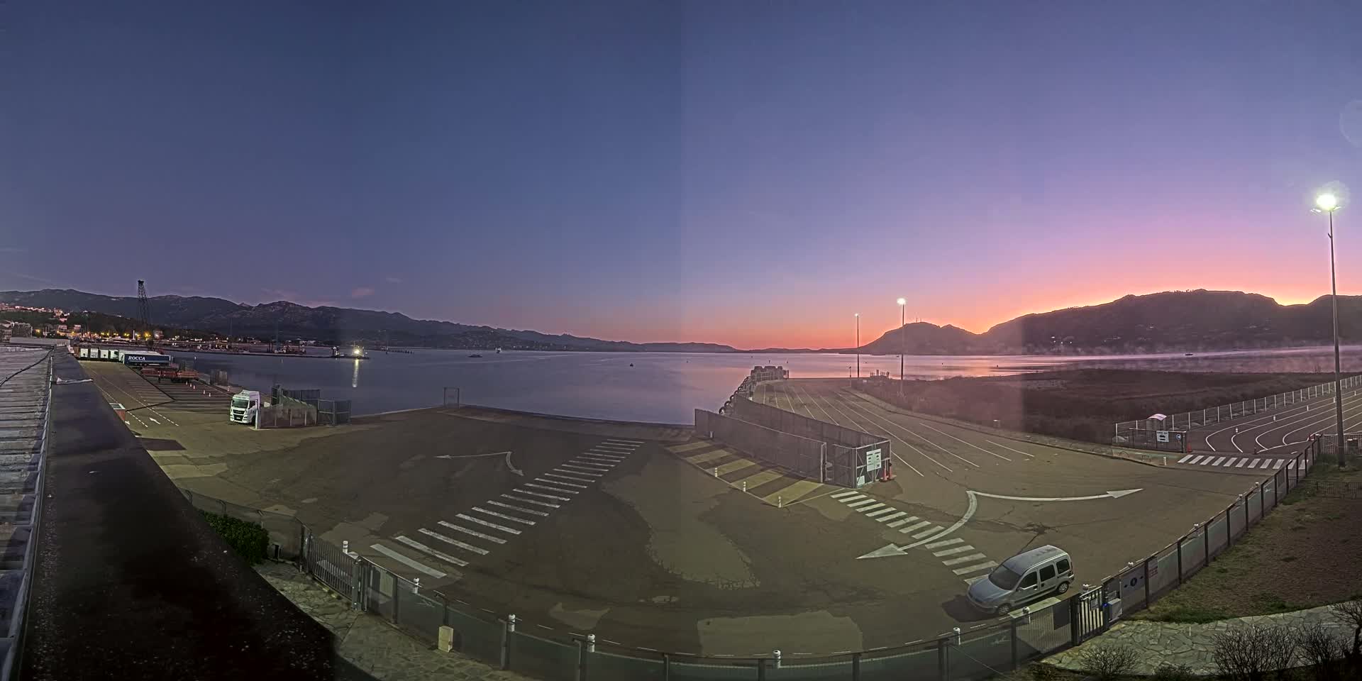 An empty port area with a few vehicles, under a twilight sky, with calm water and distant mountains in the background.