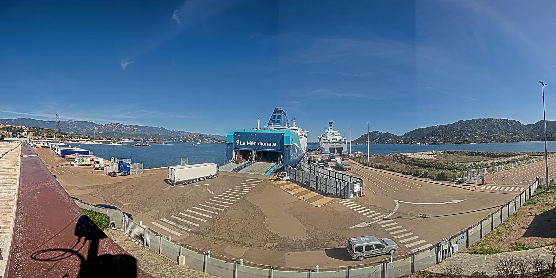 A ferry is docked at a port on a sunny day, with trucks and cars surrounding it and mountains in the background.
