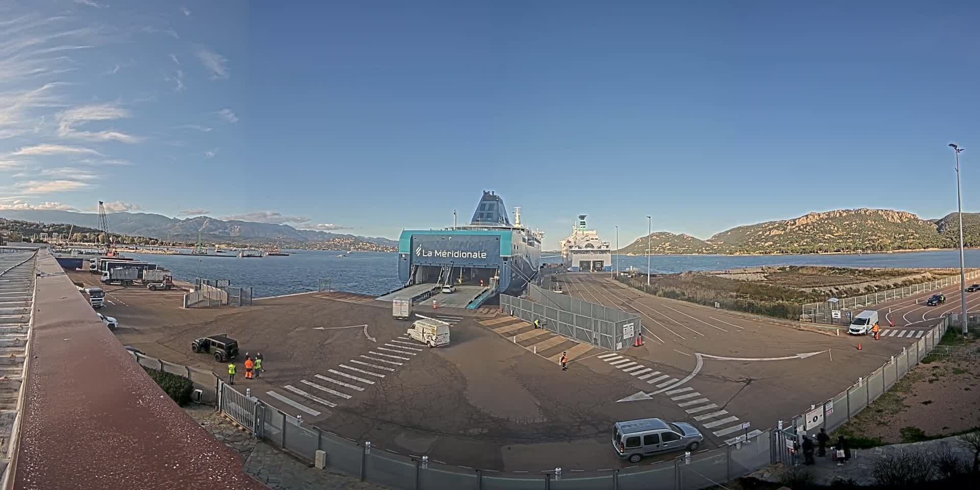 A large ferry is docked at a port on a sunny day with mountains visible in the distance.