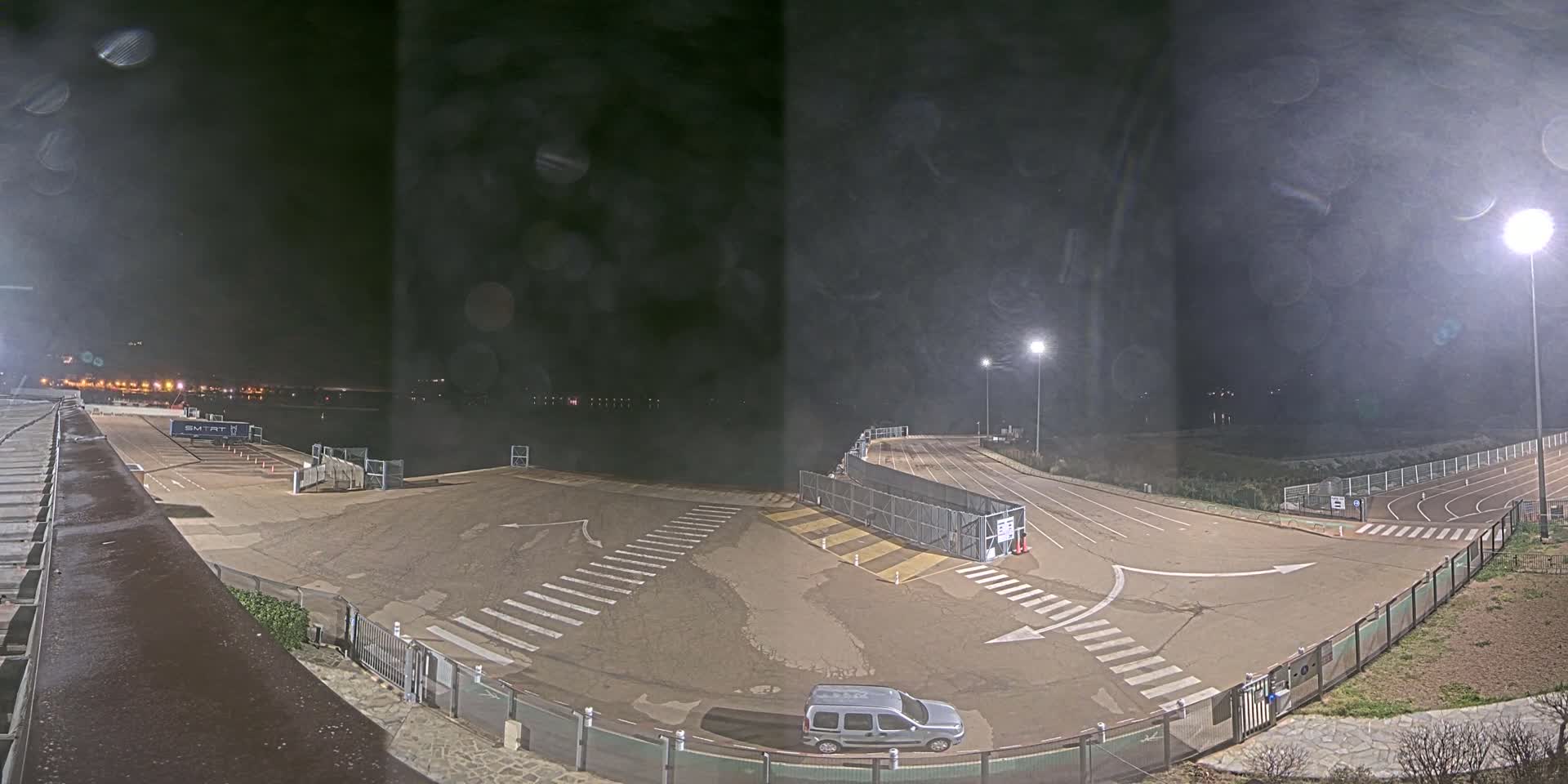 An empty, paved area with crosswalks and a running track is illuminated at night under a dark, cloudy sky, with a single vehicle parked near the center.