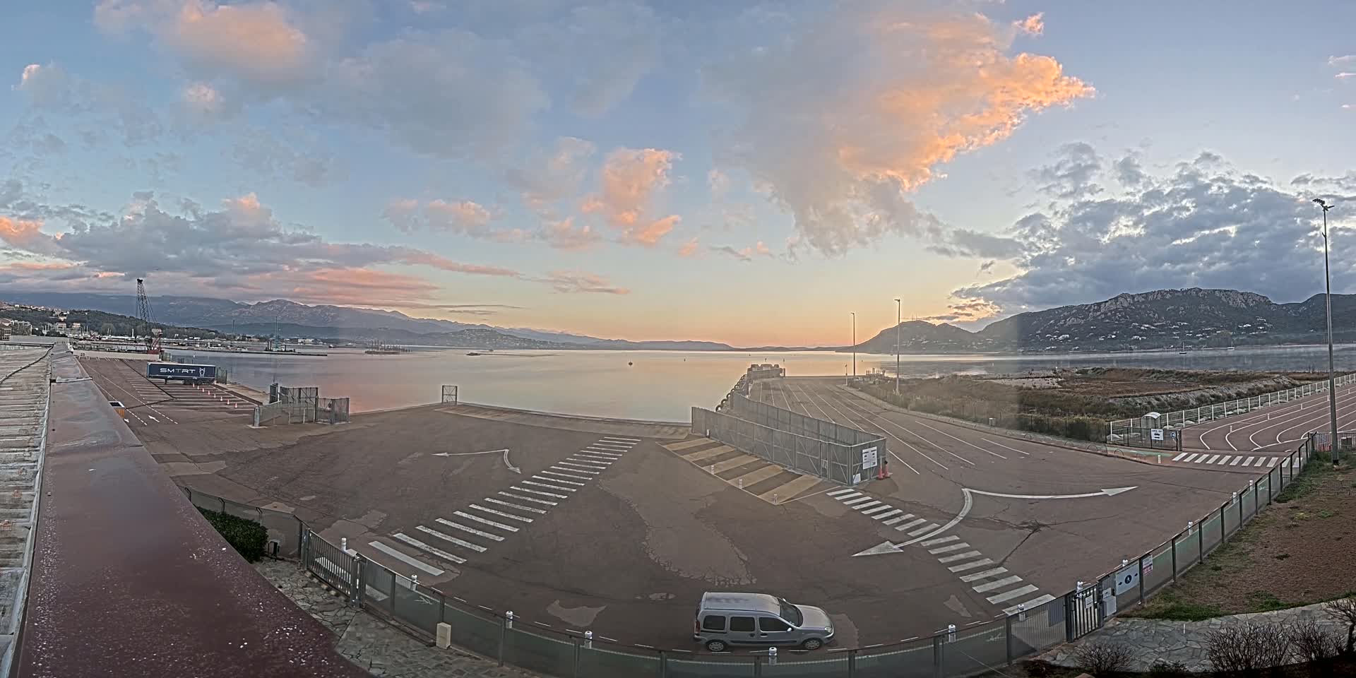 A mostly empty harbor area with a calm body of water, mountains in the background, and a pinkish sunrise sky.