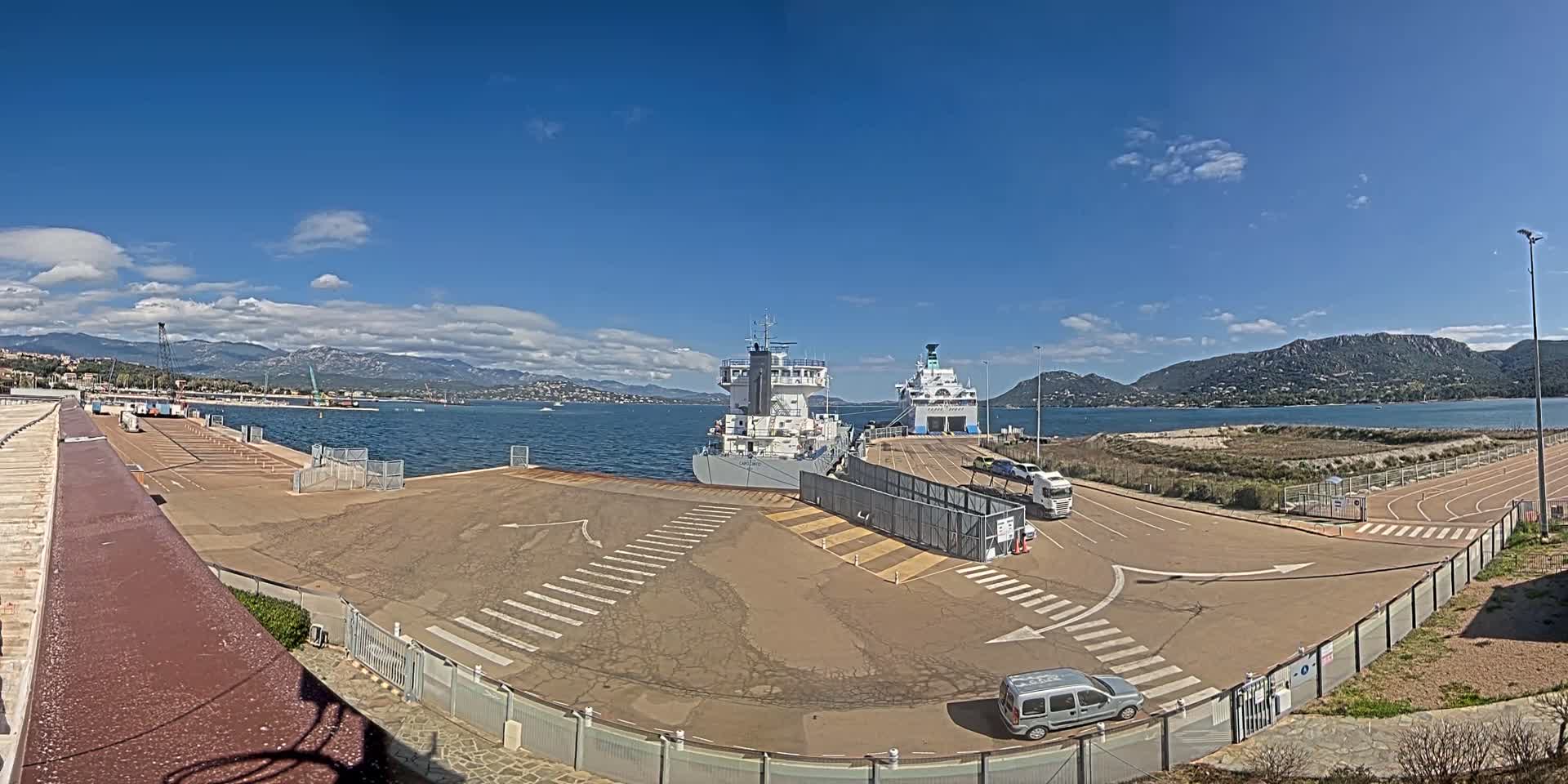 Two ships are docked at a port on a sunny day with partly cloudy skies and mountains in the distance.