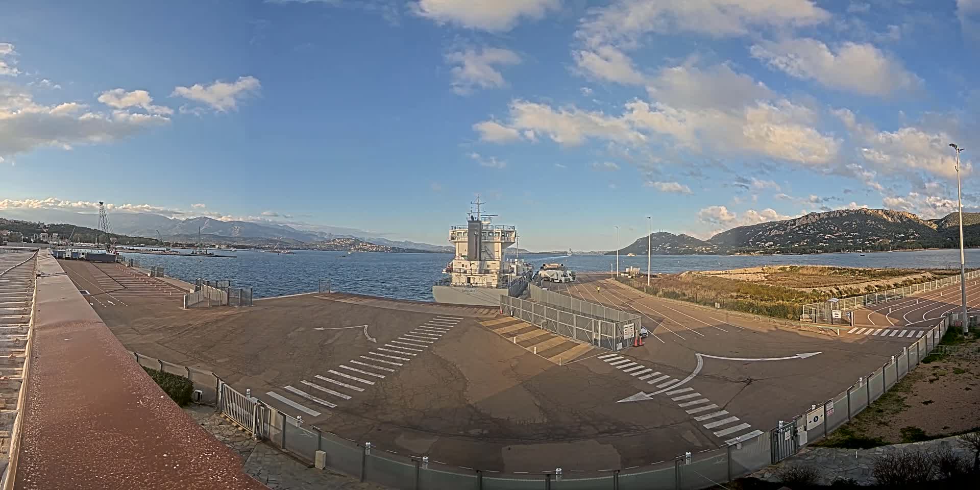 A large cargo ship is docked at a port under a partly cloudy sky, with mountains visible in the distance.