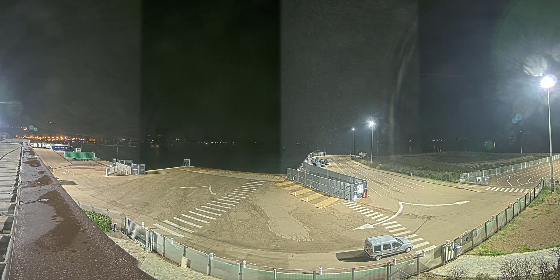 At night, an empty ferry terminal with a single vehicle is illuminated by bright lights, overlooking a dark body of water.