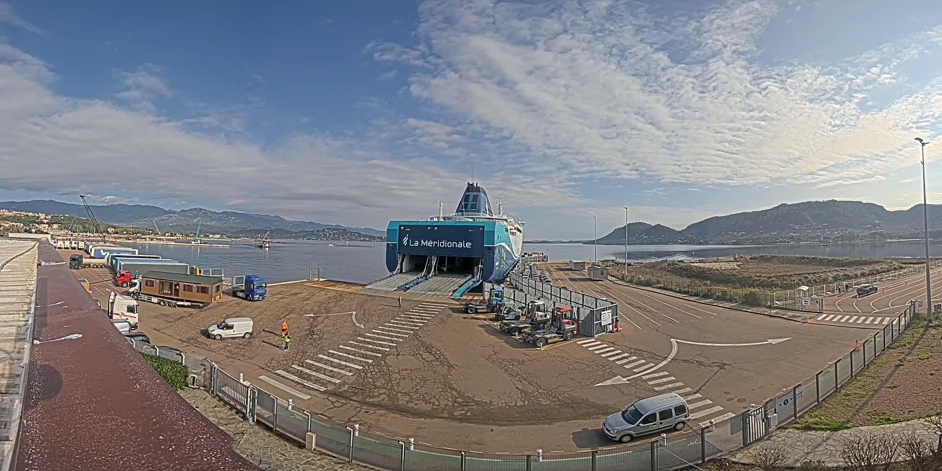 A large ferry is docked at a port under a partly cloudy sky, with several trucks and other vehicles nearby.