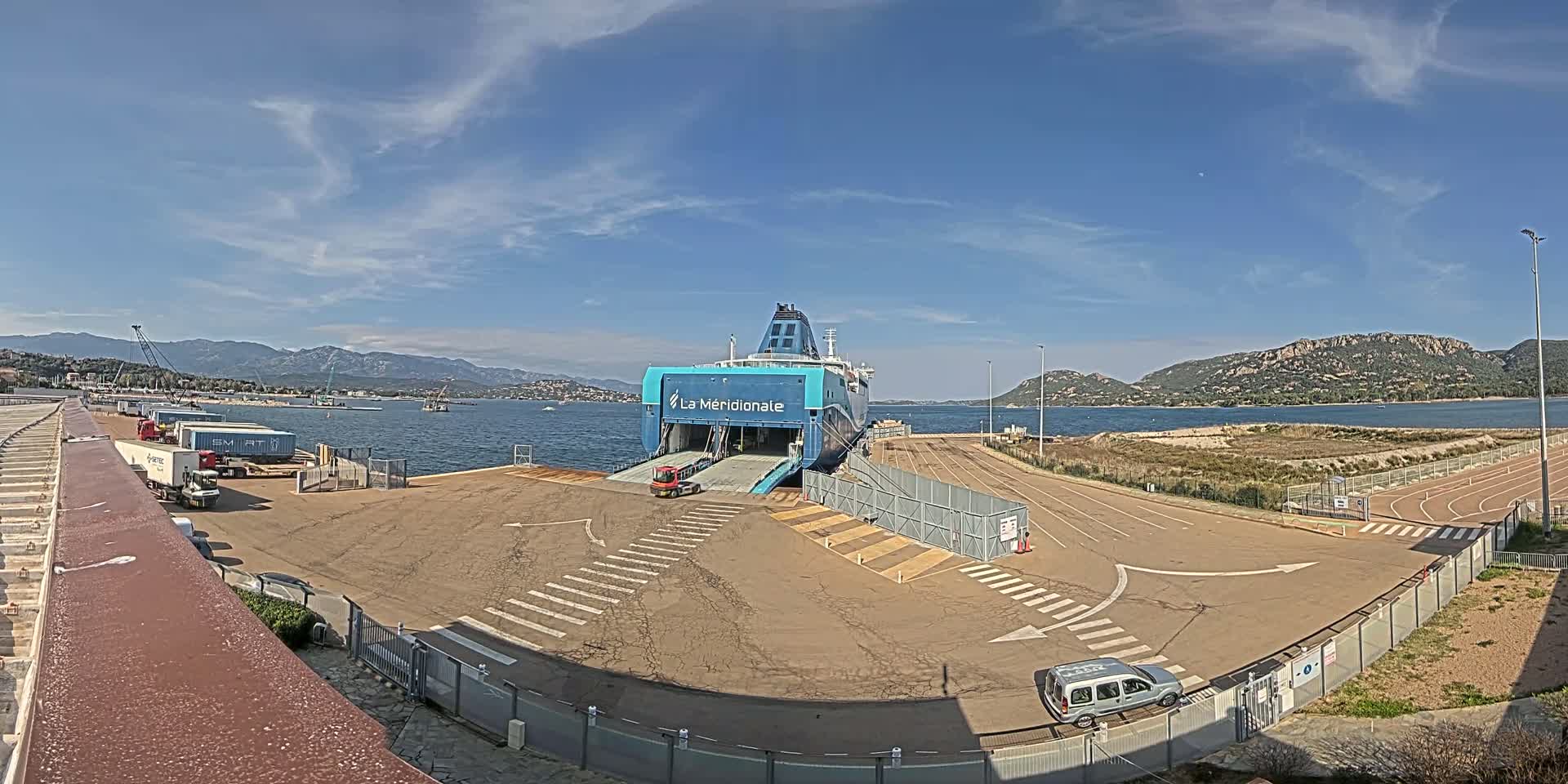A large ferry is docked at a port on a sunny day, with mountains visible in the distance and several trucks parked nearby.