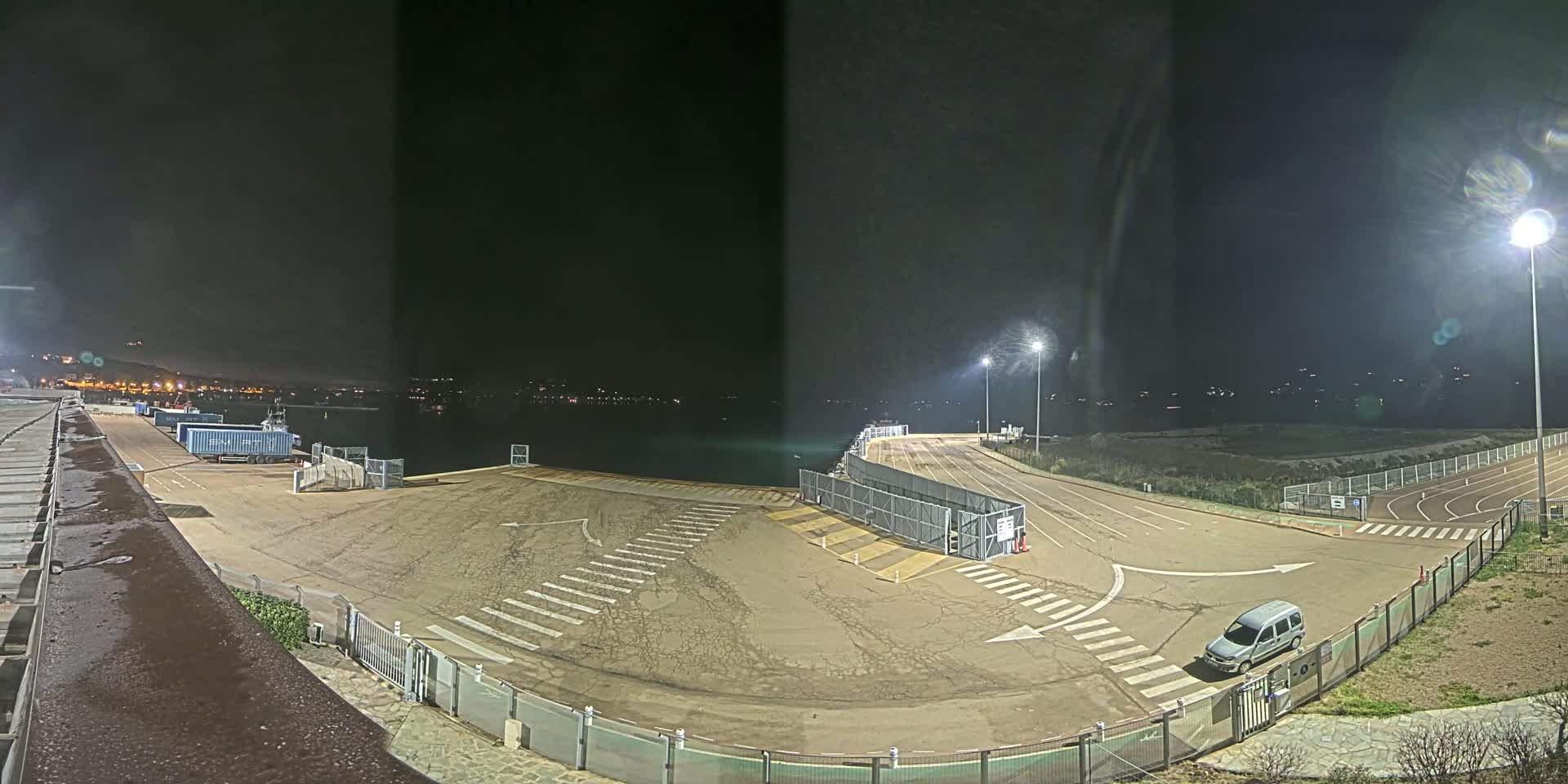 An empty, well-lit nighttime port area with a single vehicle, bordered by fences and featuring crosswalks and several shipping containers.