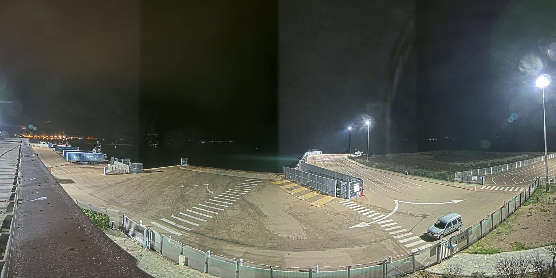 A nighttime view of an empty ferry terminal with a single vehicle parked near a gated entrance, under artificial light.