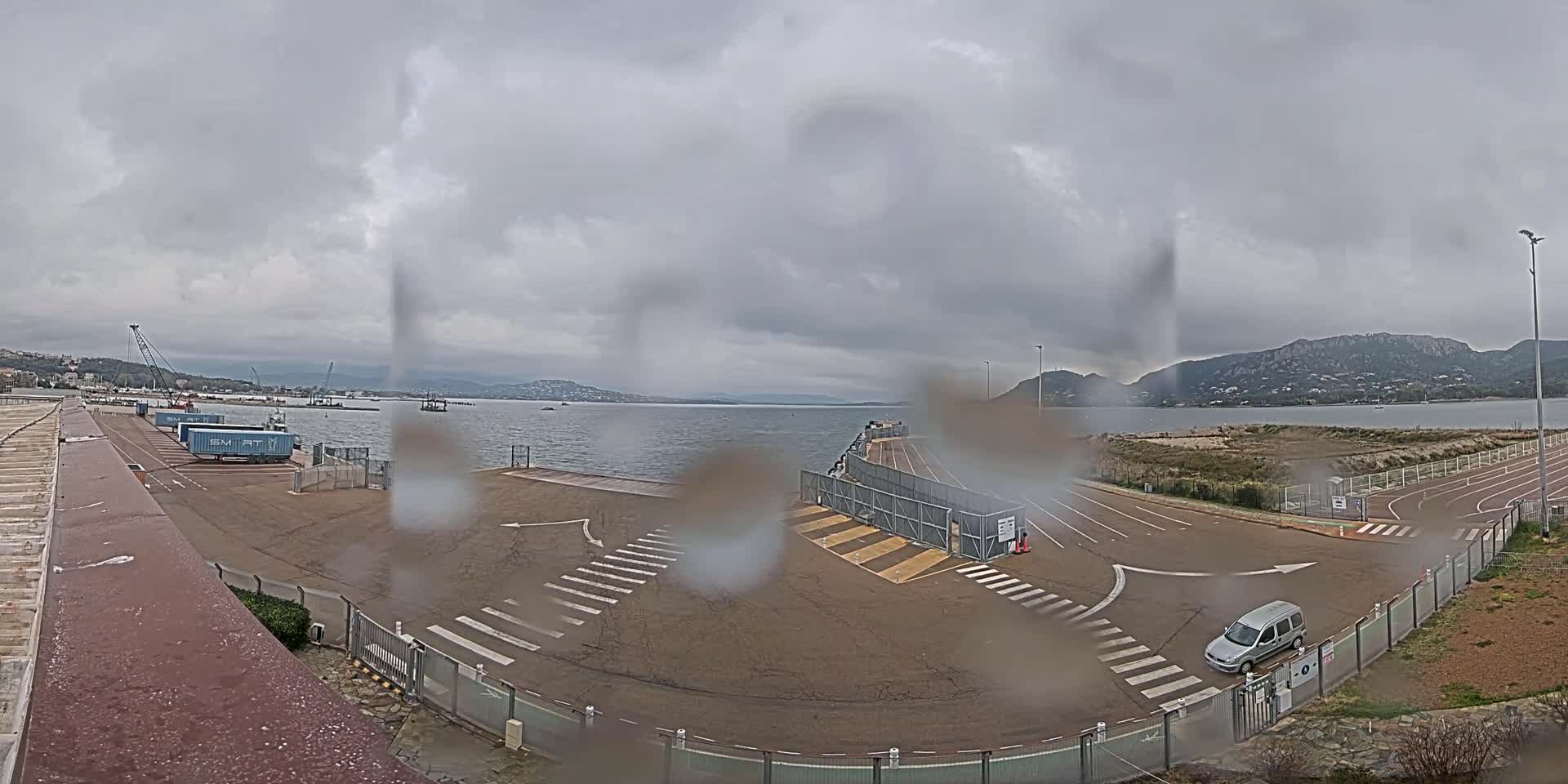 A rain-covered view of a port area with a calm body of water, mountains in the distance, and a single vehicle parked near a gate.