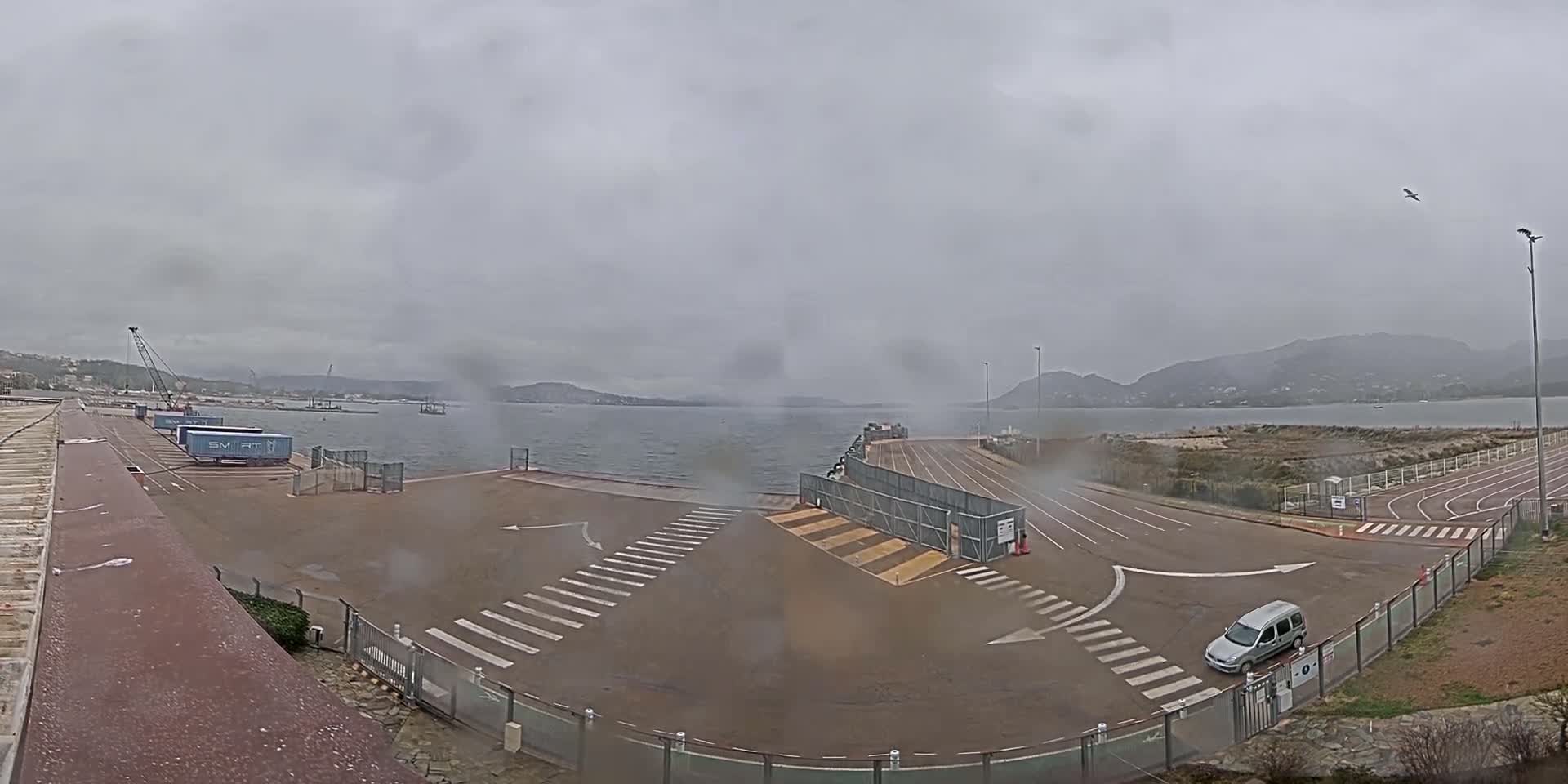 A mostly empty port area with a single car visible, under an overcast sky, overlooks a calm body of water and distant hills.