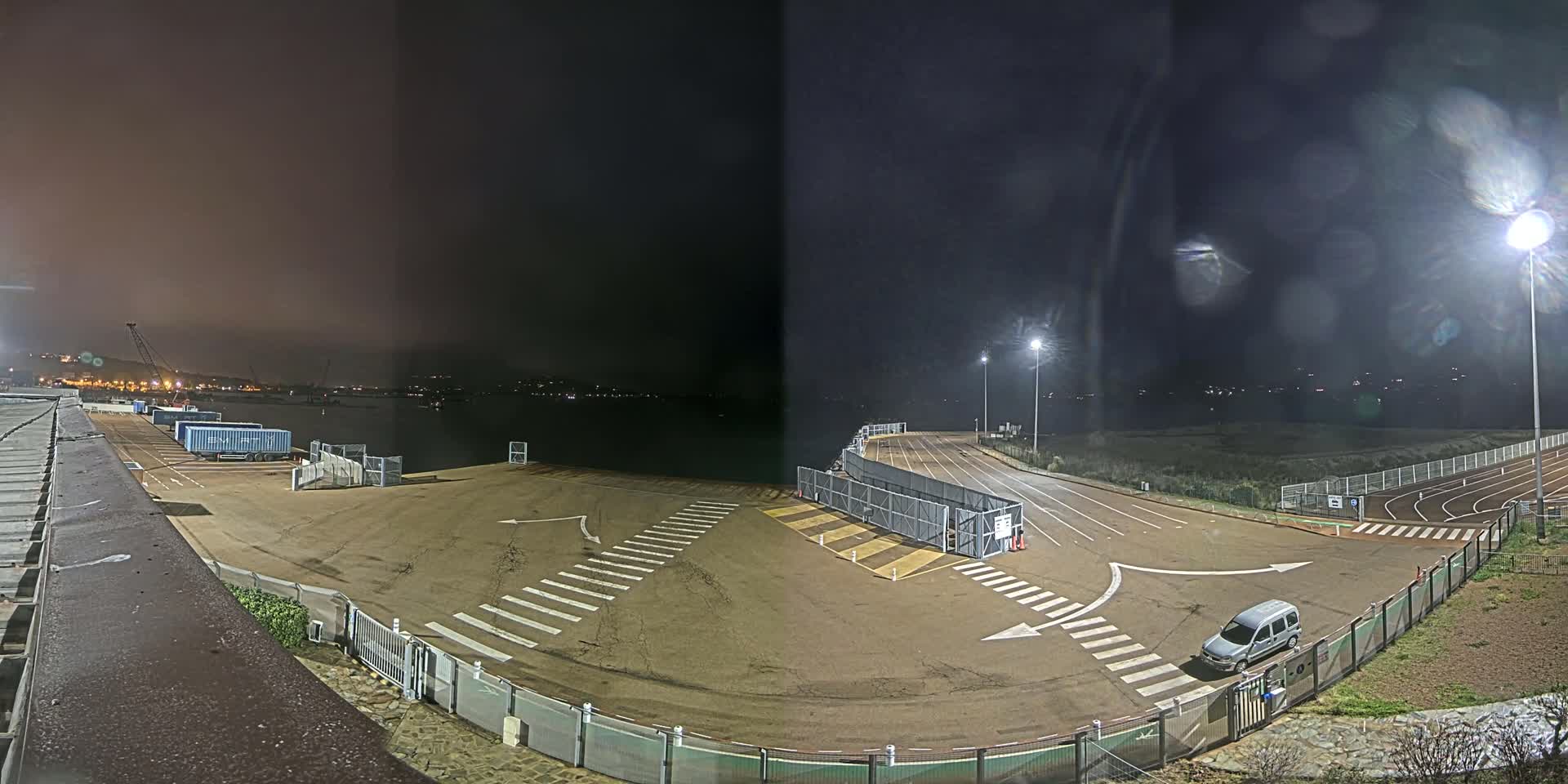 A nighttime view of an empty port area with a single van, a roadway, and a running track, under a dark, slightly hazy sky.