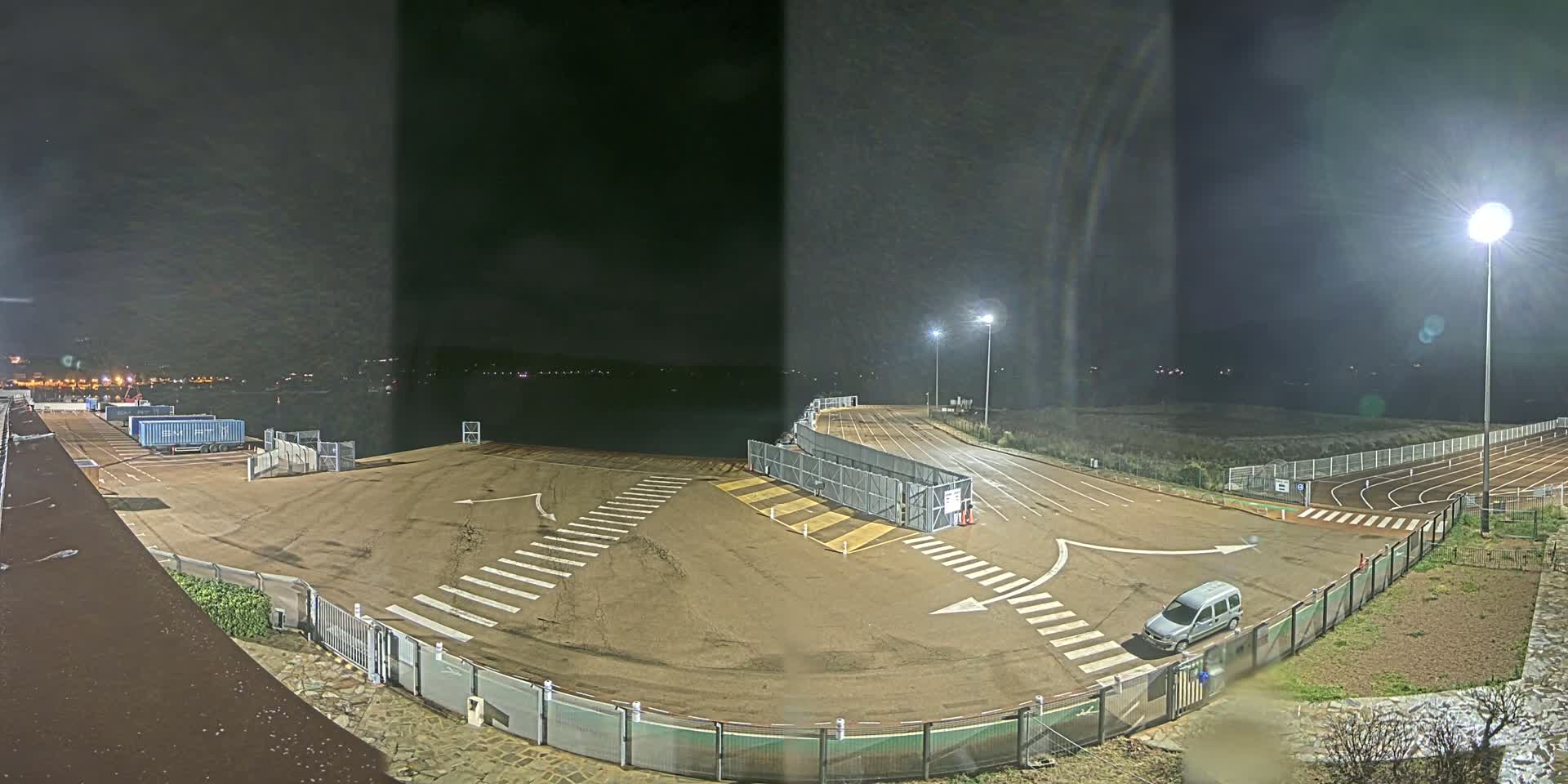 A nighttime view of an empty, paved area with crosswalks, fencing, and a single van, next to a running track, under bright artificial lights.