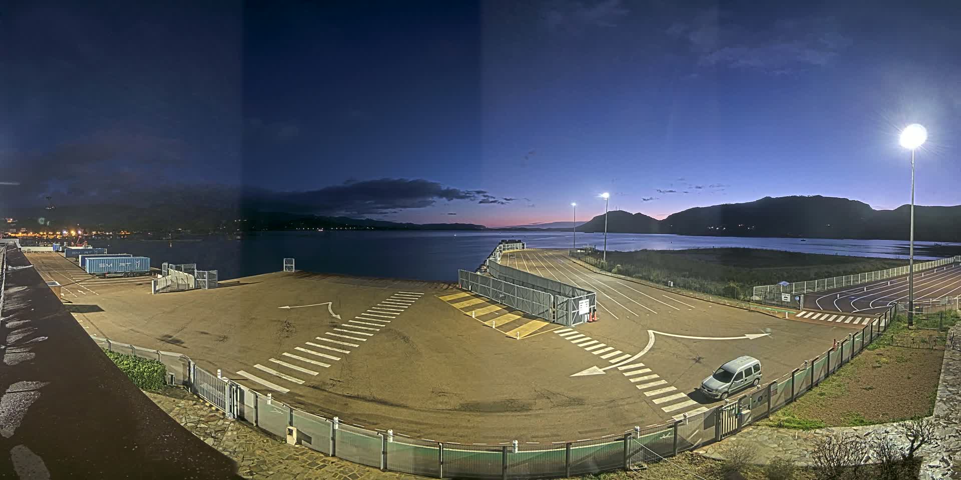 An empty ferry terminal at twilight, under artificial lights, overlooks a calm body of water surrounded by hills.