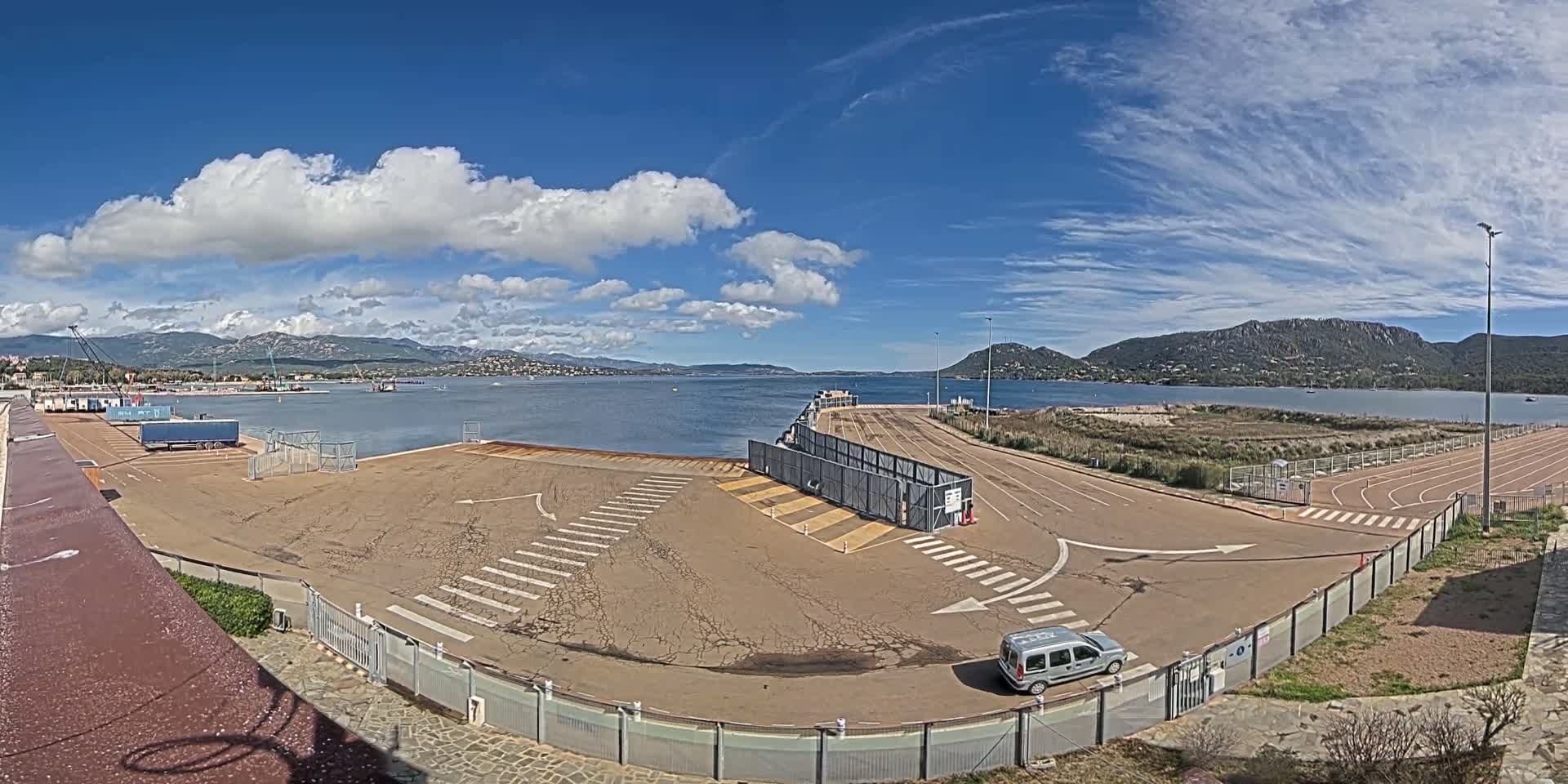 A mostly sunny day shows a mostly empty port area with a single car parked near a gated entrance, overlooking a calm body of water surrounded by hills and mountains.