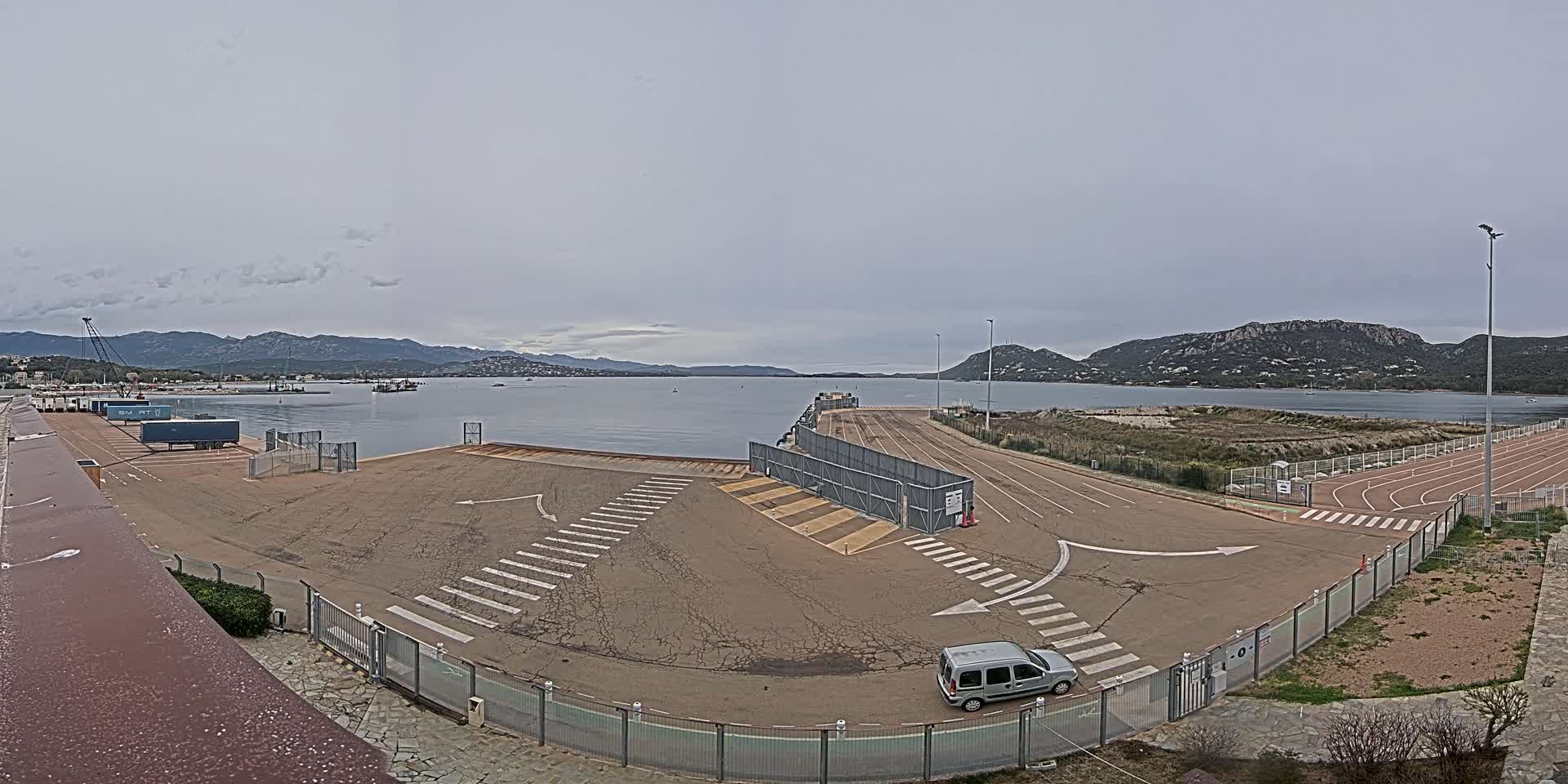 A mostly empty port area with a single vehicle, under an overcast sky, overlooks a calm body of water surrounded by mountains.