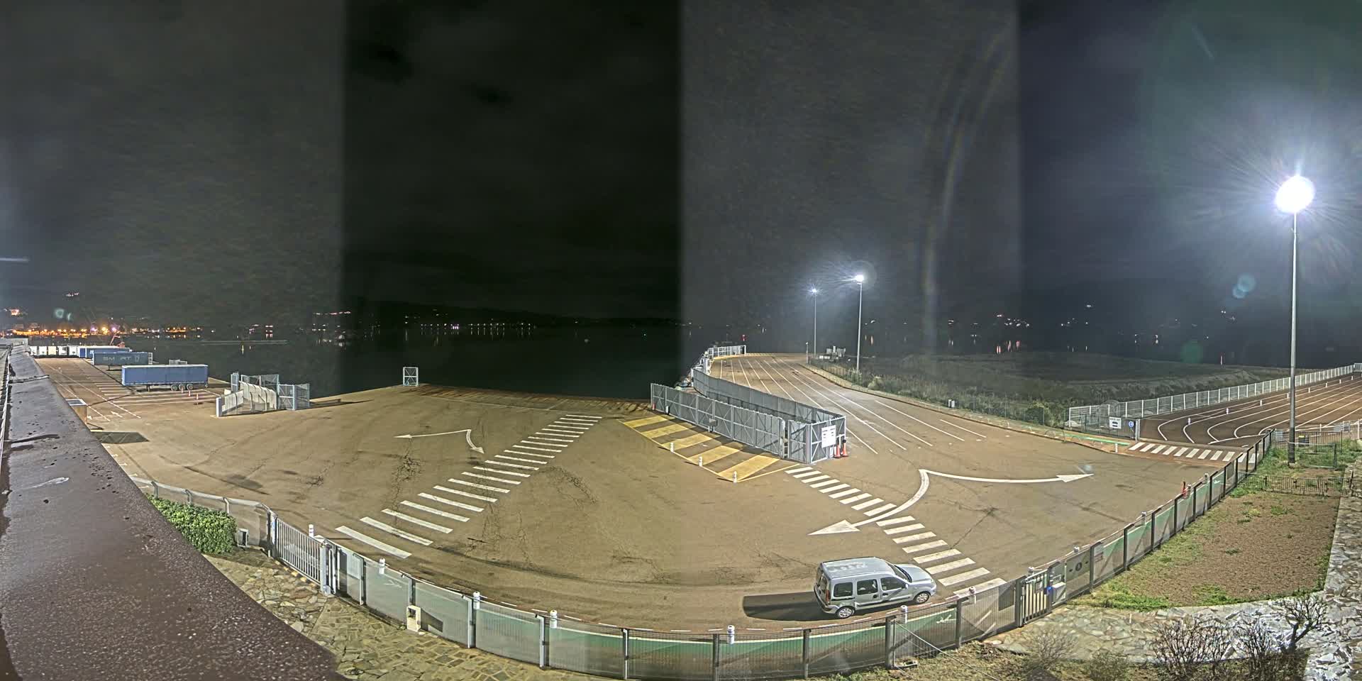 An empty, well-lit nighttime parking lot with a single van, bordered by a fence and overlooking a calm body of water.