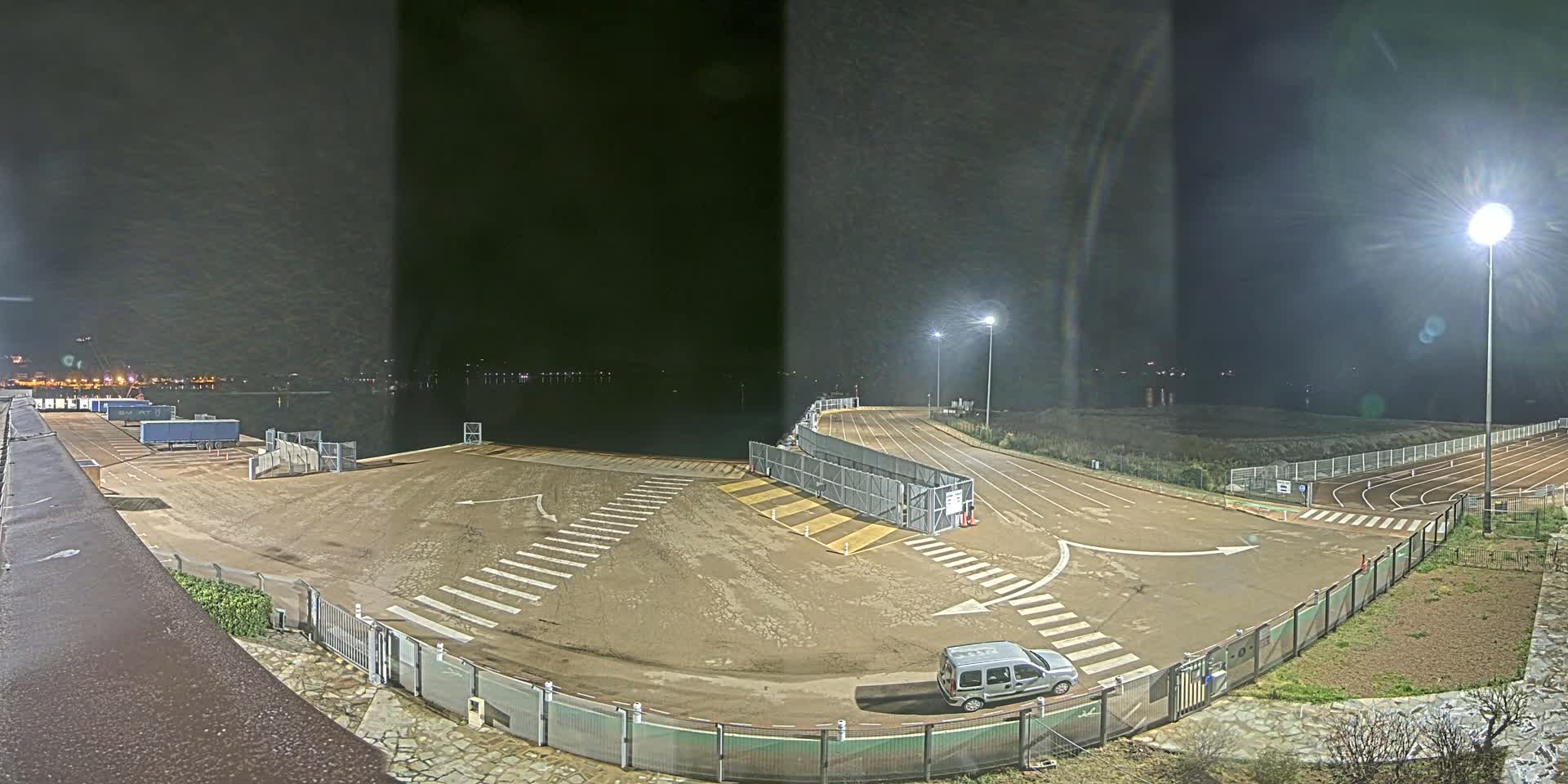 A nighttime, outdoor view of a mostly empty port area with a vehicle parked near a fenced-in section, adjacent to a running track.
