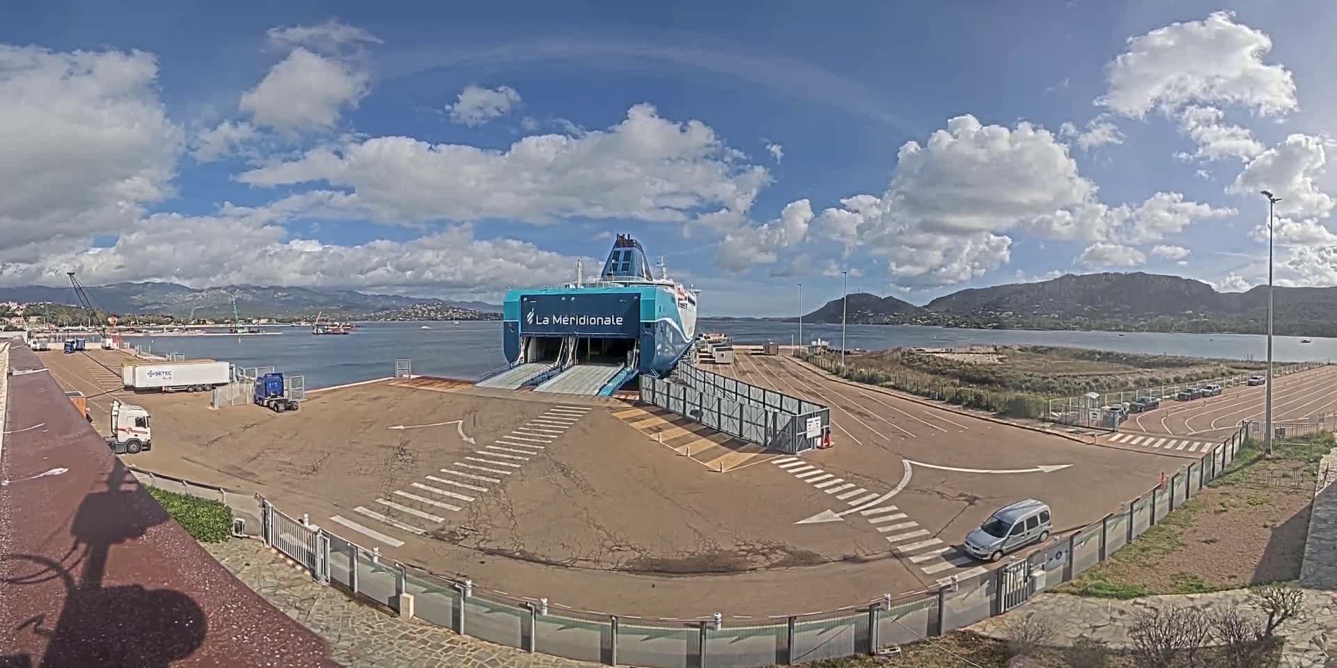 A large ferry is docked at a port on a partly cloudy day, with several vehicles parked nearby and mountains visible in the distance.