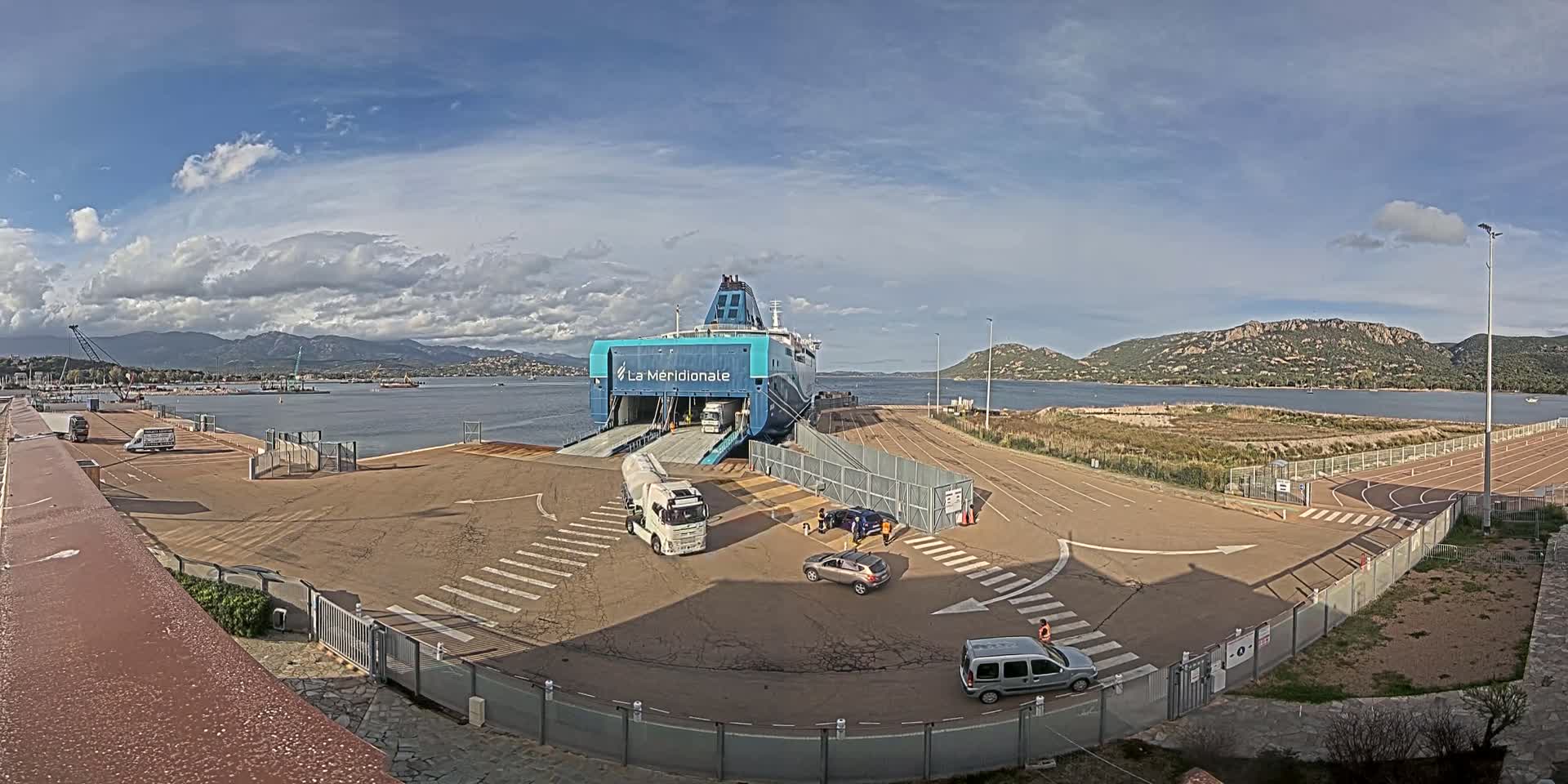 A large ferry is docked at a port, with several vehicles on the dock and in the process of loading or unloading, under a partly cloudy sky.