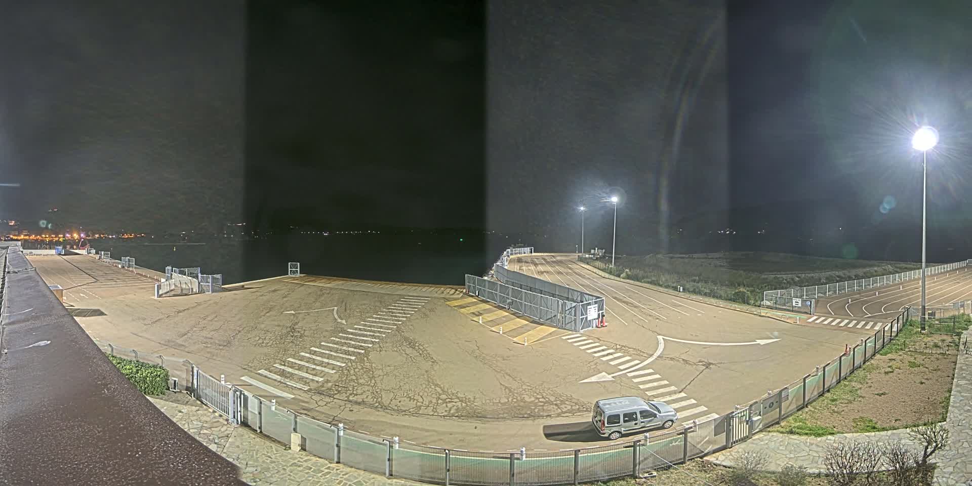 A nighttime view of an empty, paved area with lane markings, gates, and a single vehicle, overlooking a dark body of water under bright artificial lights.