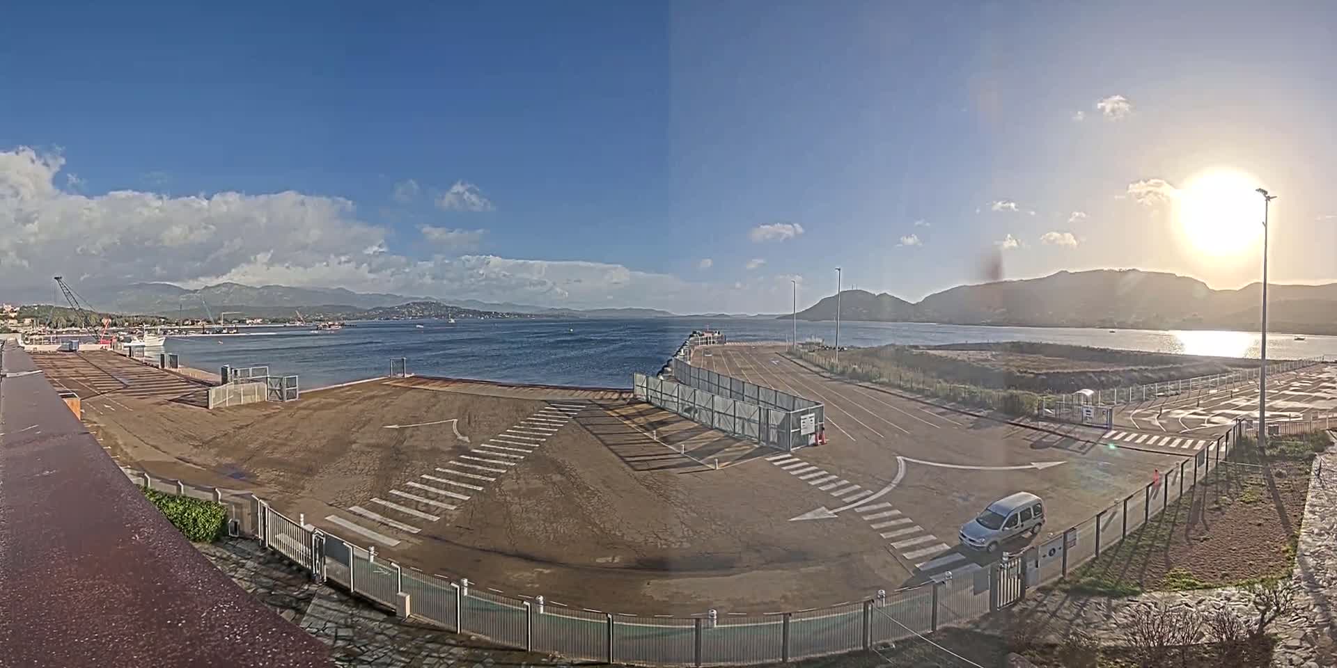 A mostly sunny day shows a mostly empty port area with a single car parked near a gated entryway, overlooking a calm body of water and distant mountains.