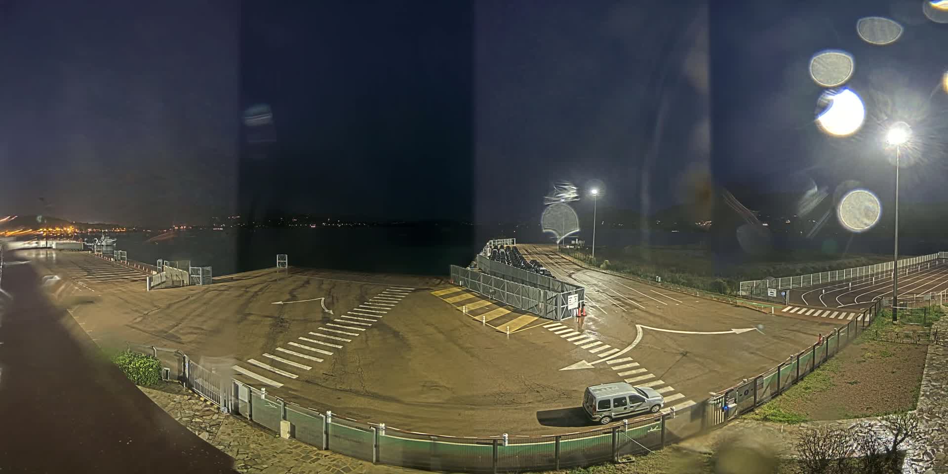 A nighttime view of an empty parking lot near a body of water, with a running track visible in the background, under slightly rainy conditions.