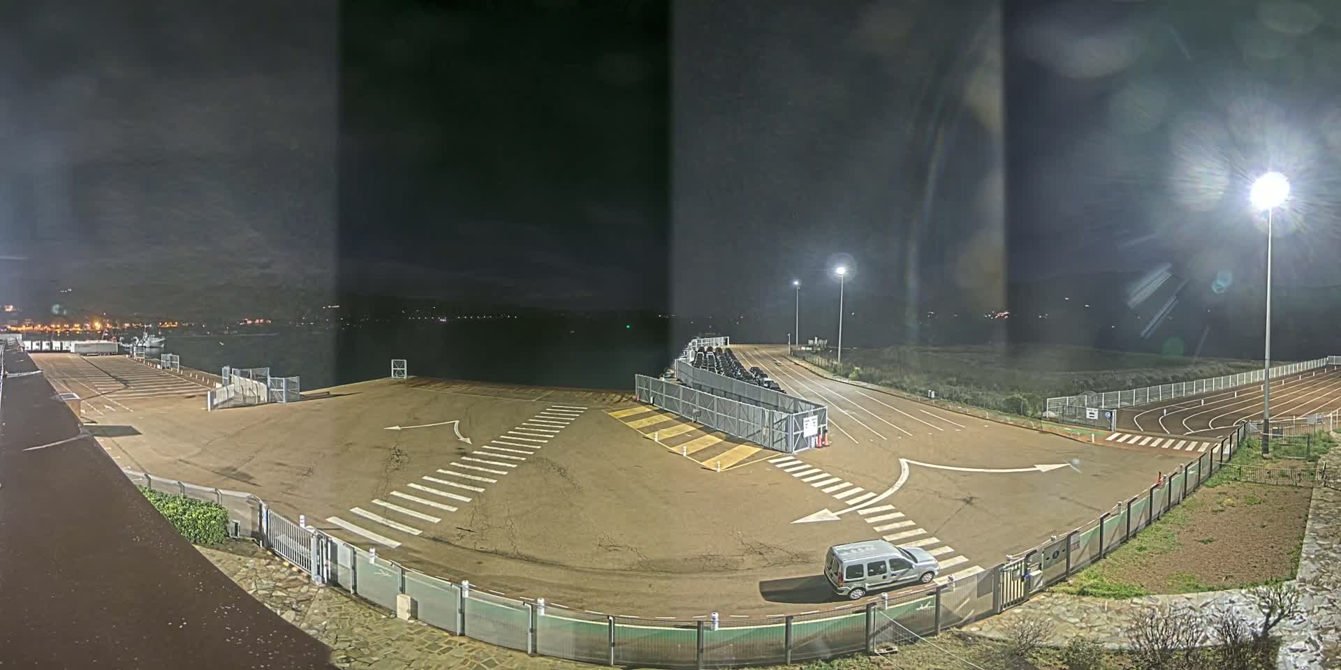 A nighttime view of an empty parking lot next to a body of water, with a row of cars parked behind a fence and a running track visible in the lower right corner.