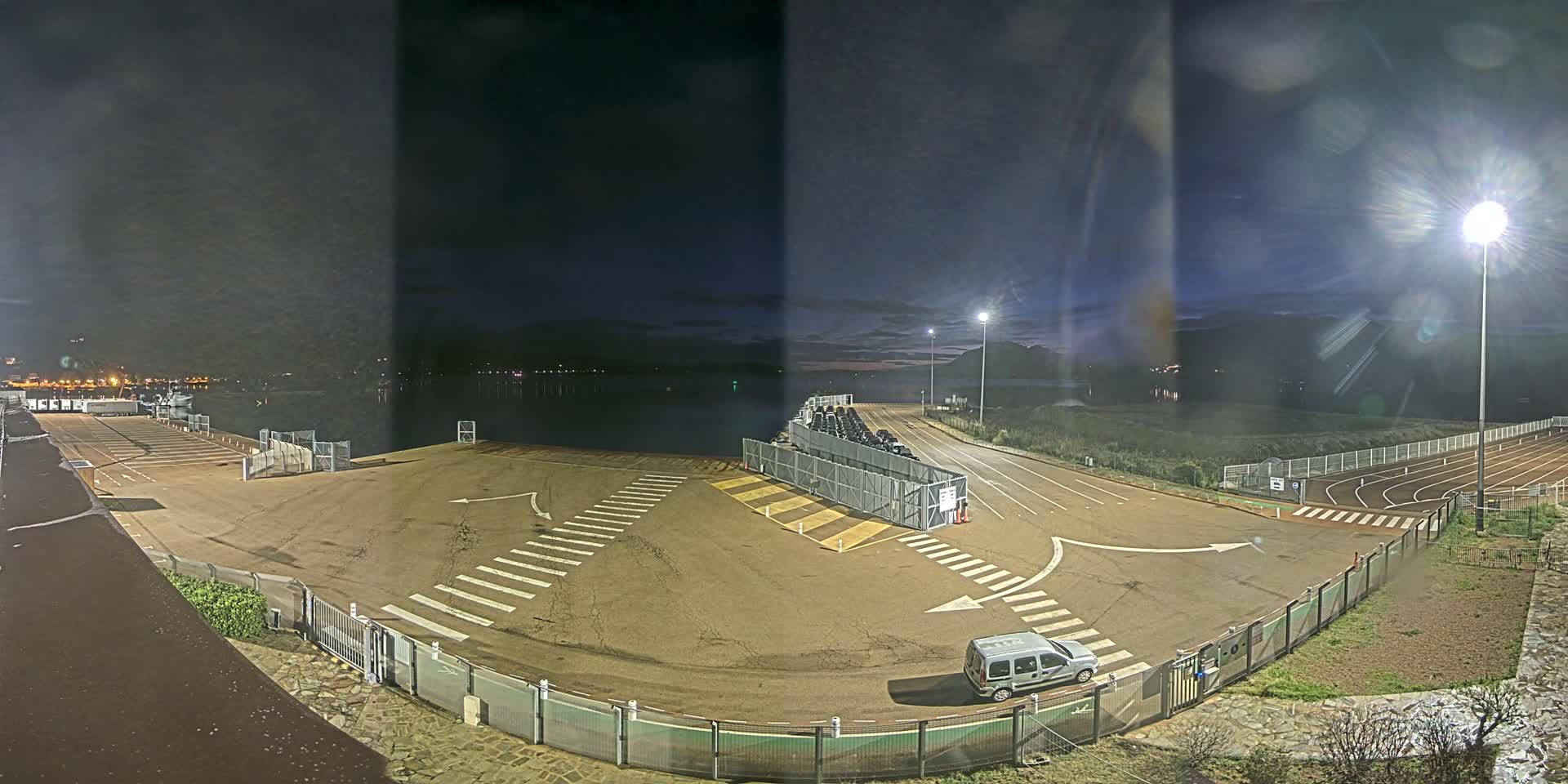 An empty, nighttime ferry terminal with a single vehicle parked near a gate, next to a body of water under a partly cloudy sky.