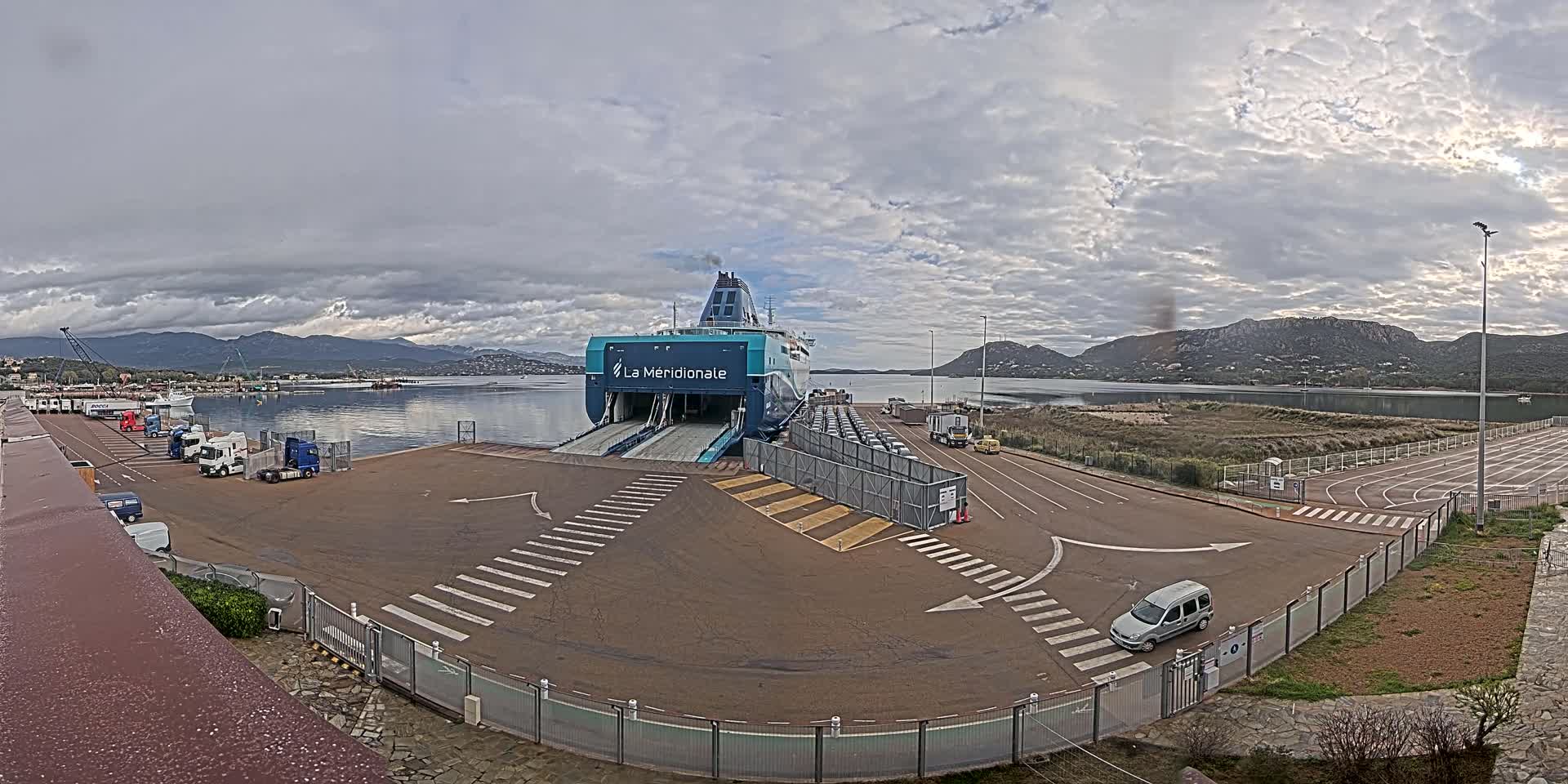 A large ferry is docked at a port under a cloudy sky, with several vehicles parked nearby and mountains visible in the distance.