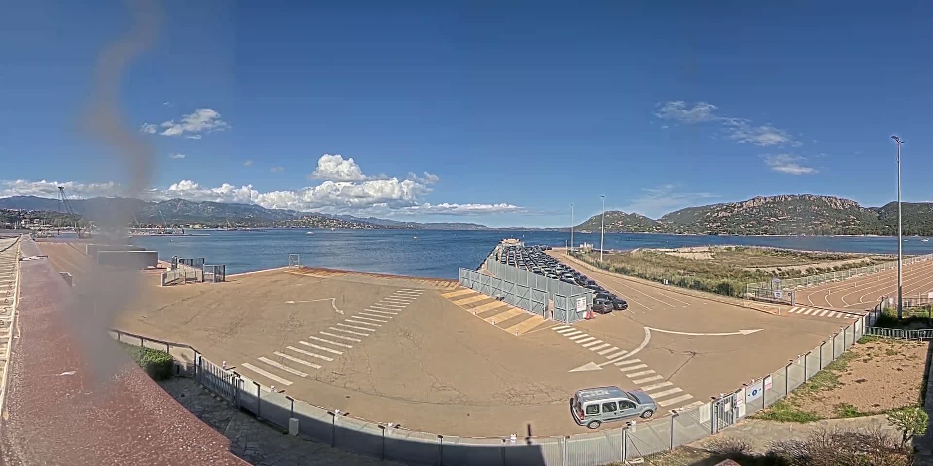 A mostly sunny day shows a harbor area with a parking lot, a boat launch, and a calm body of water with mountains in the background.
