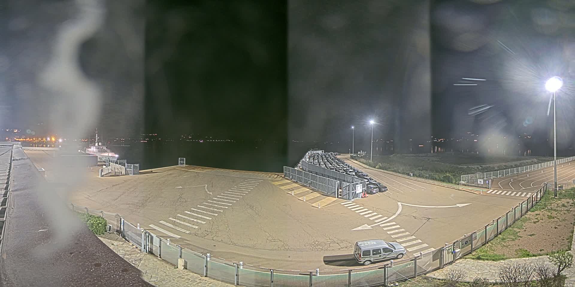 A nighttime view of a port area with numerous cars parked behind a fence, a single van in a nearby lot, and a ship visible in the distance across the water.
