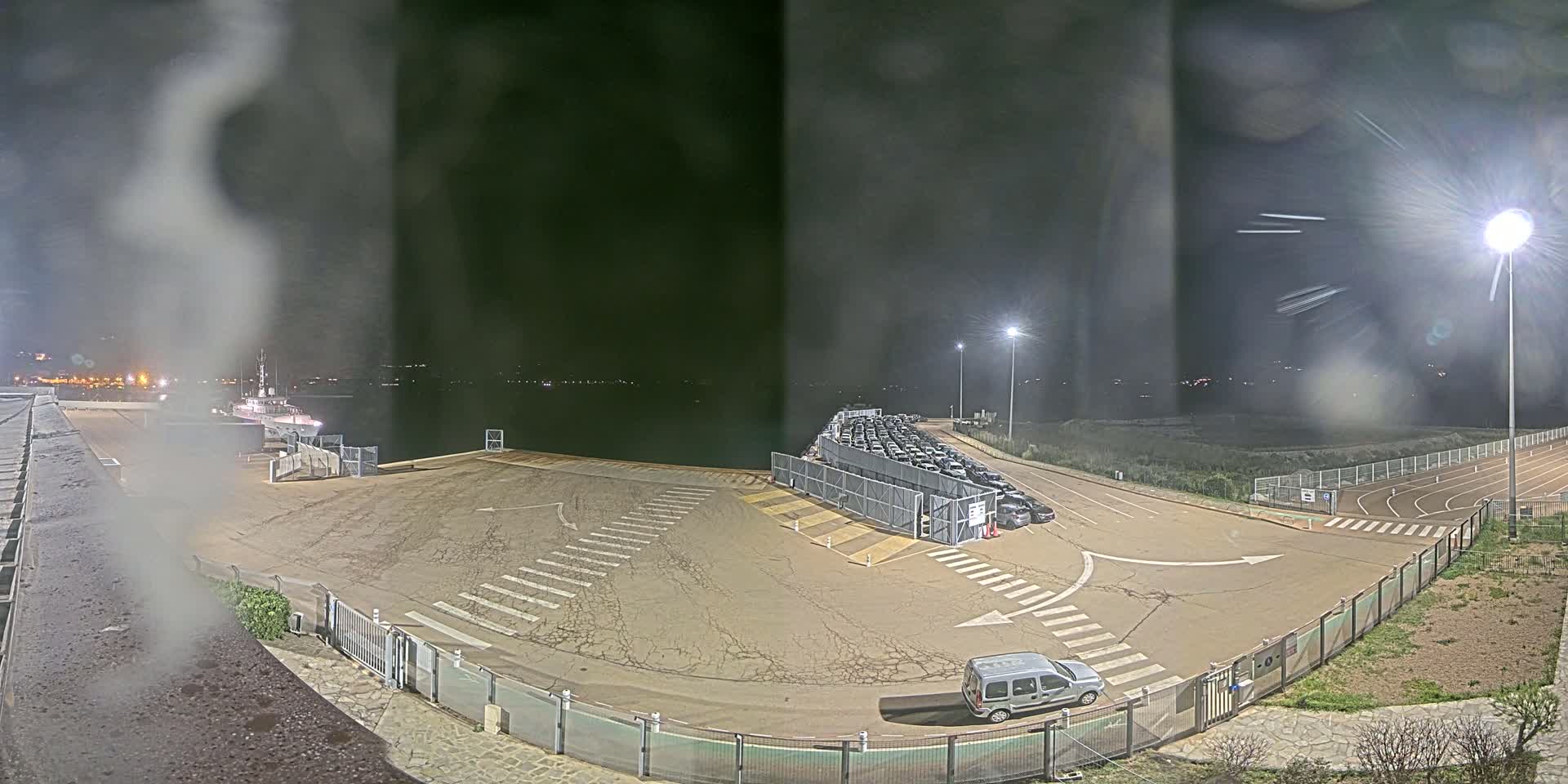 A nighttime view of an empty port area with a ship docked, a row of parked cars behind a fence, and a single vehicle in a nearby lot, under cloudy skies.